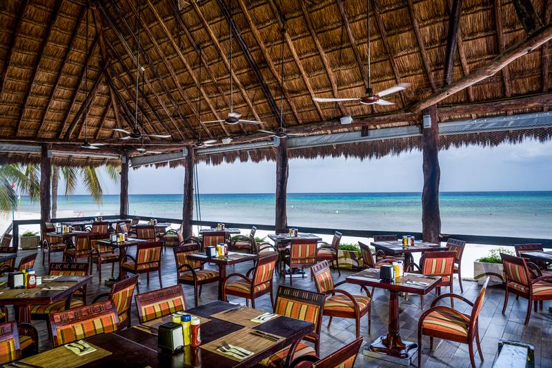 Beachfront spa treatment room with two massage tables, ocean view through open wooden structure, and thatched-roof bungalows visible beyond palm trees.