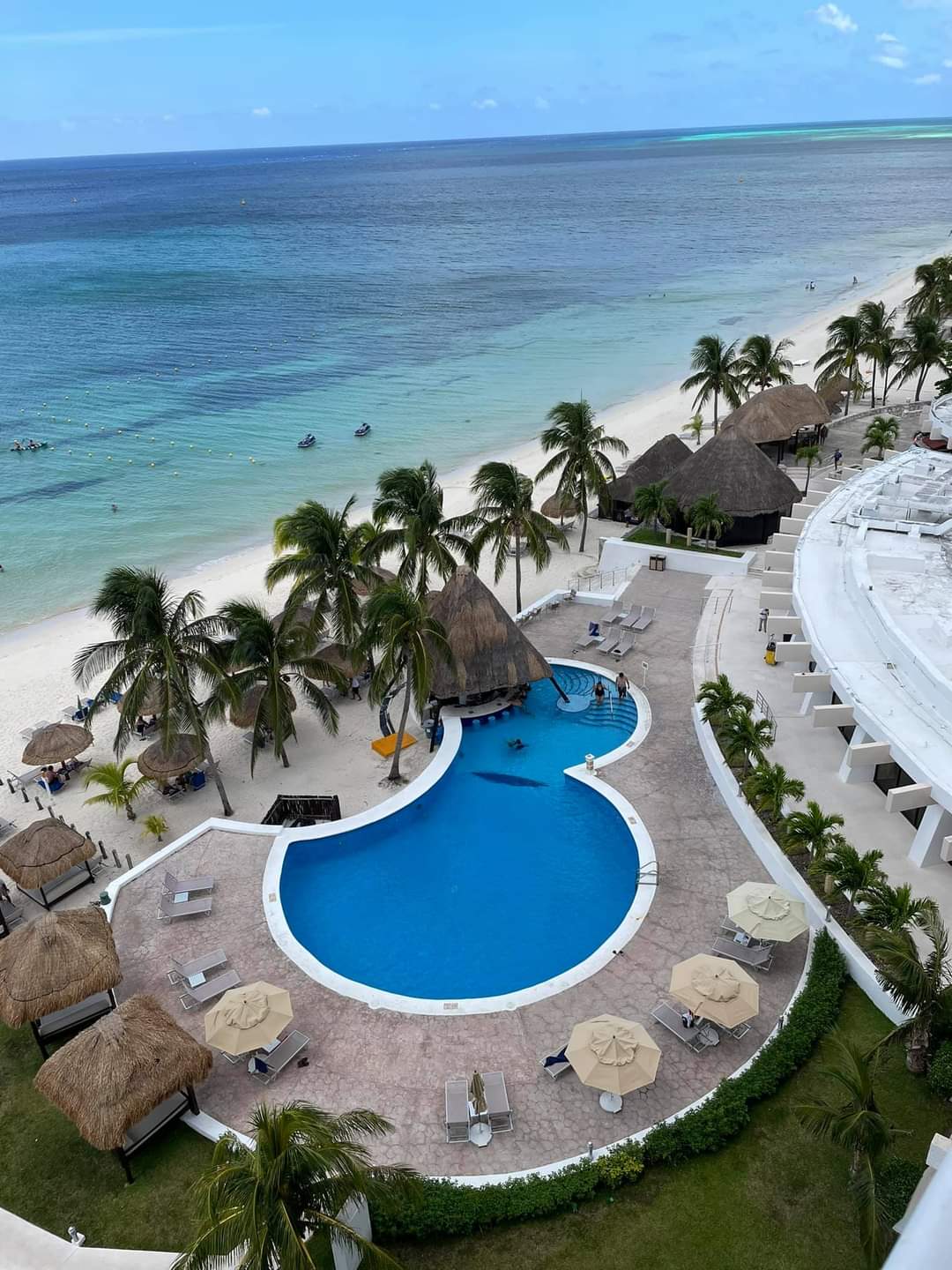 Resort pool with thatched-roof palapa, palm trees, white buildings, and ocean view under clear blue sky.