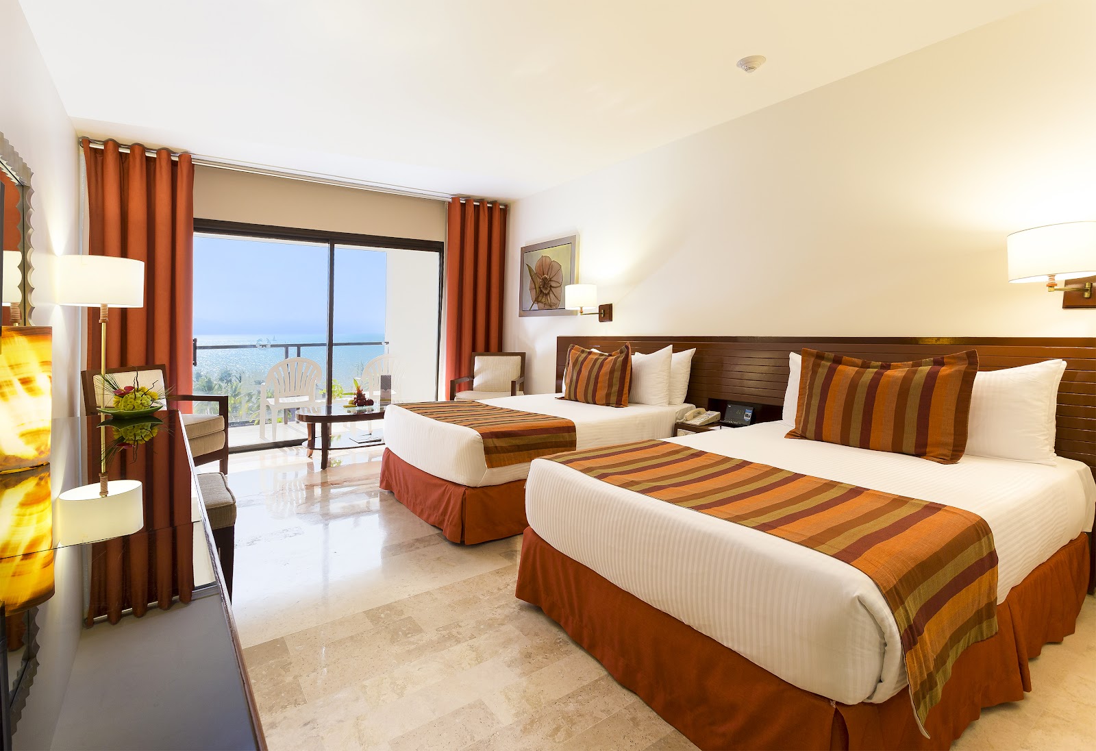 Resort room with two beds, orange striped bedding, and a balcony overlooking the ocean and coastline.