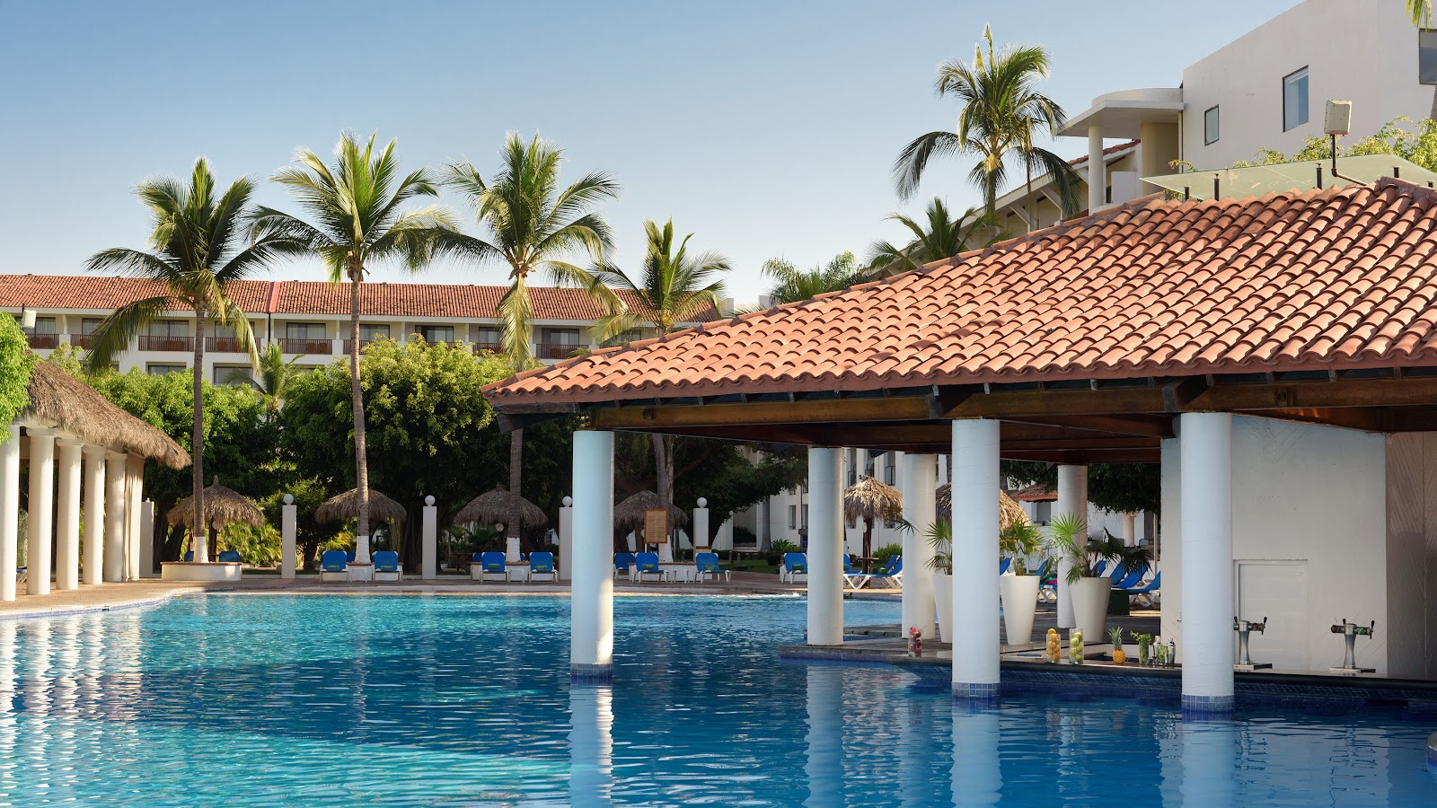 Resort pool with white columns and terracotta roof structure, palm trees, and guests on blue loungers under thatched palapas.