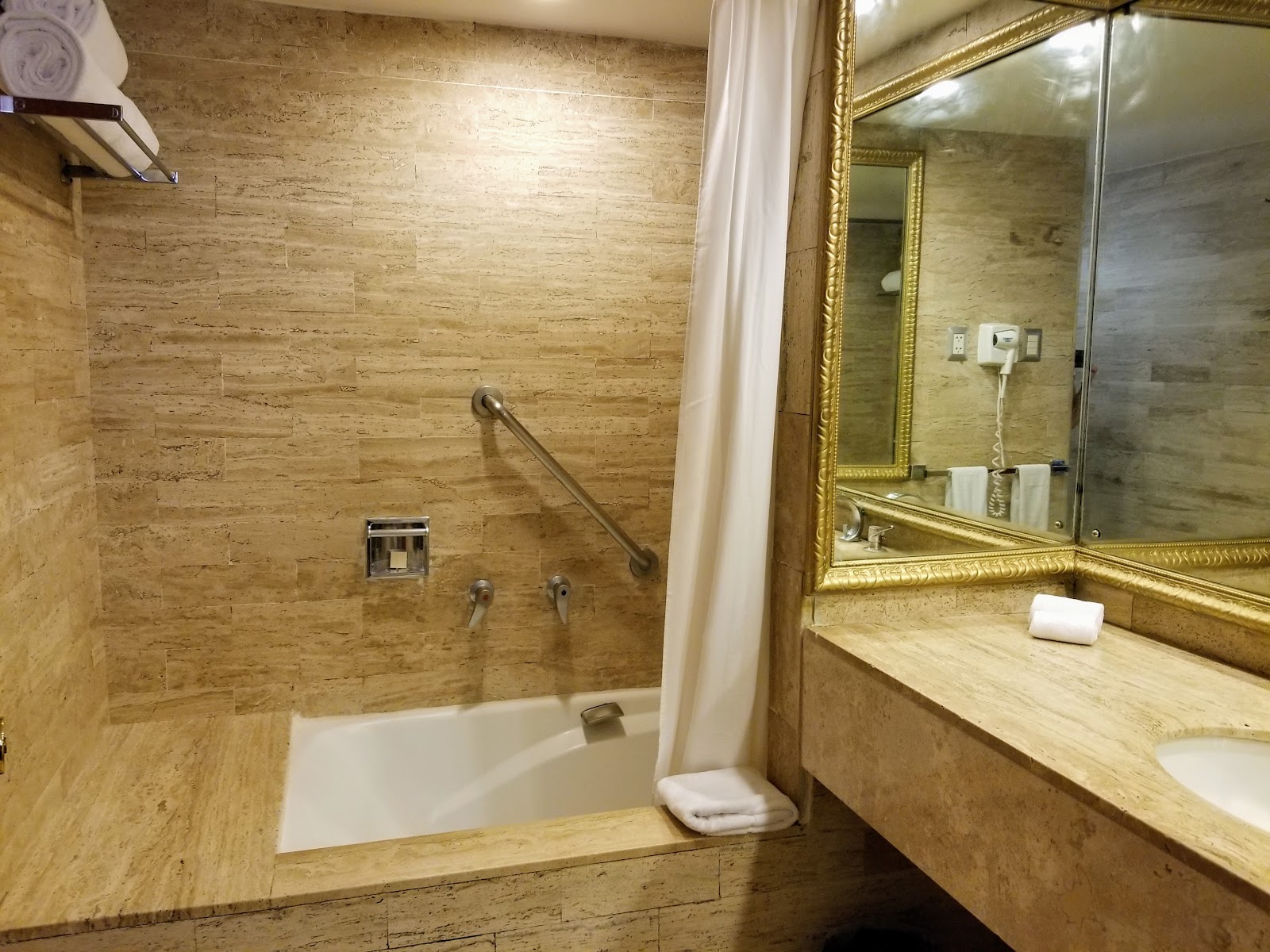 Luxurious bathroom with travertine walls, gold-framed mirror, marble vanity with pink lilies, and walk-in shower with curtain at Meliá Puerto Vallarta.