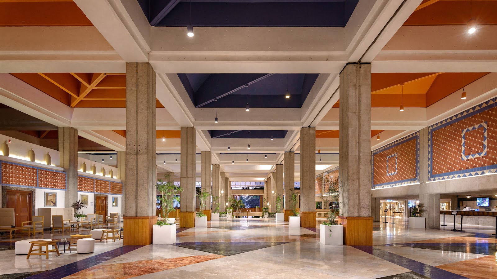 Spacious resort lobby with polished marble floors, concrete columns, orange and navy ceiling details, and decorative textile panels on the walls.