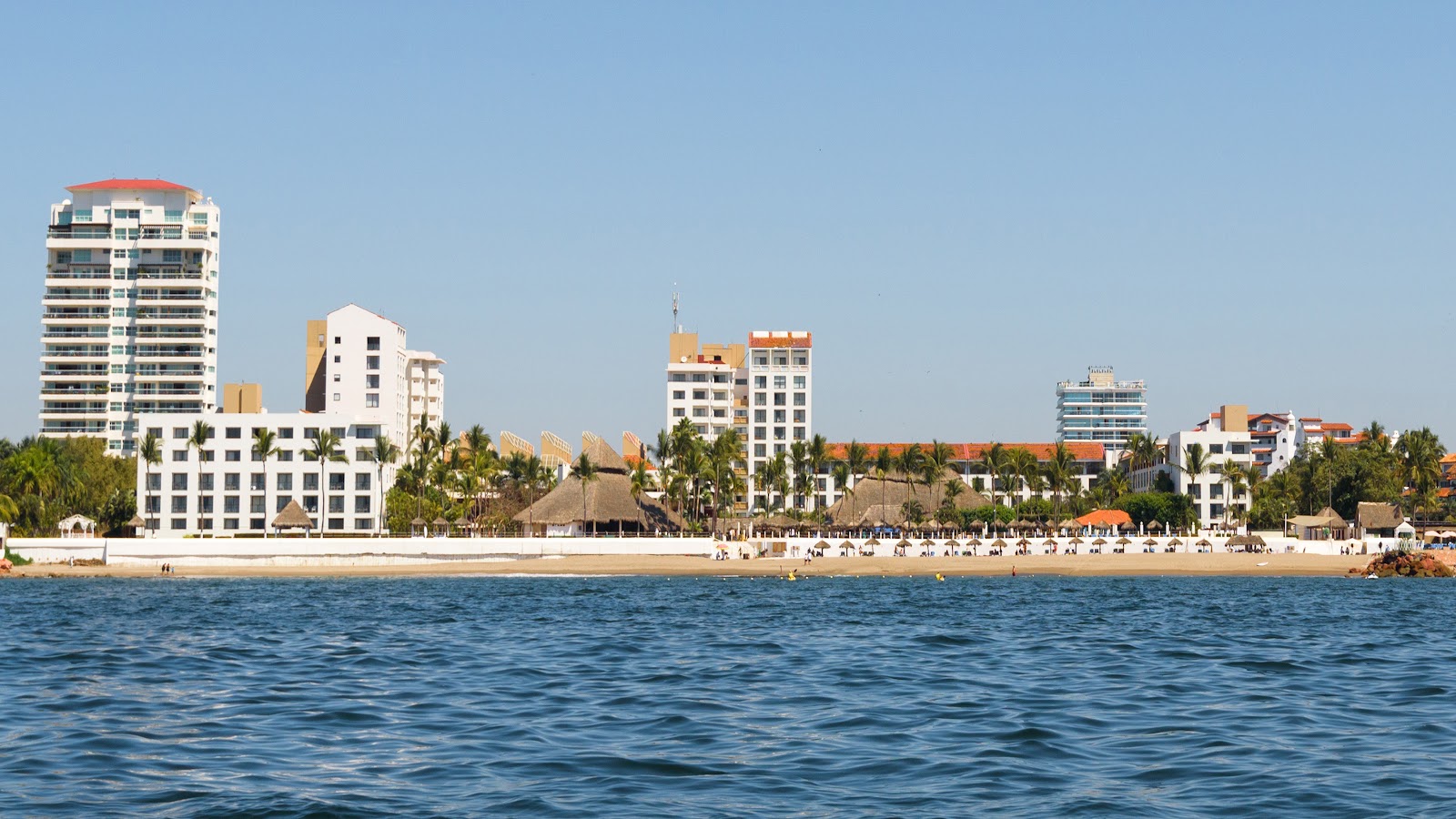 Beachfront resort buildings with white and gold facades line a sandy beach, viewed from the ocean with palm trees and loungers visible along the shore.