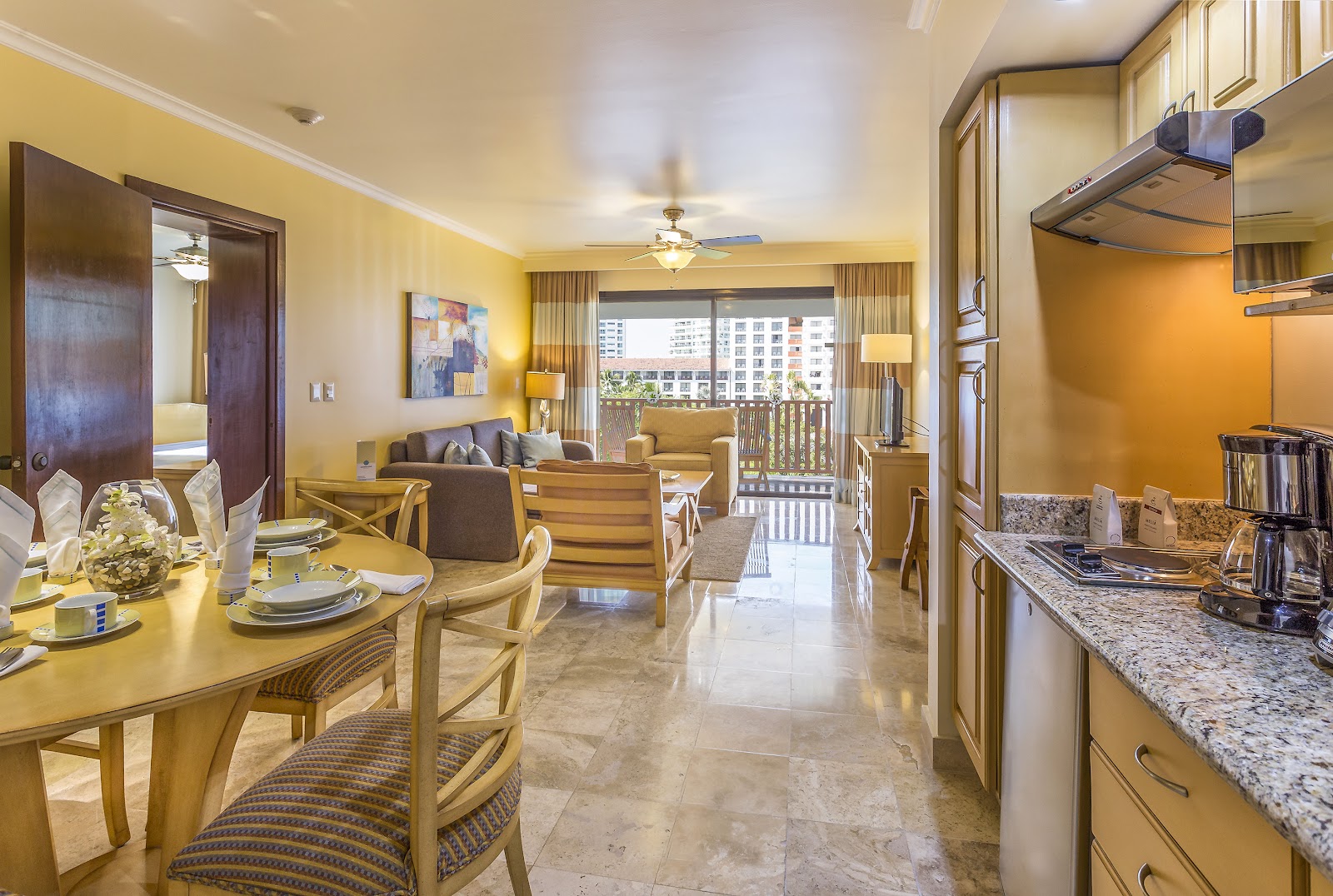 Modern dining hall with wood-slat ceiling, buffet counter displaying fresh fruits and dishes, and tables set with place settings and yellow accent chairs.
