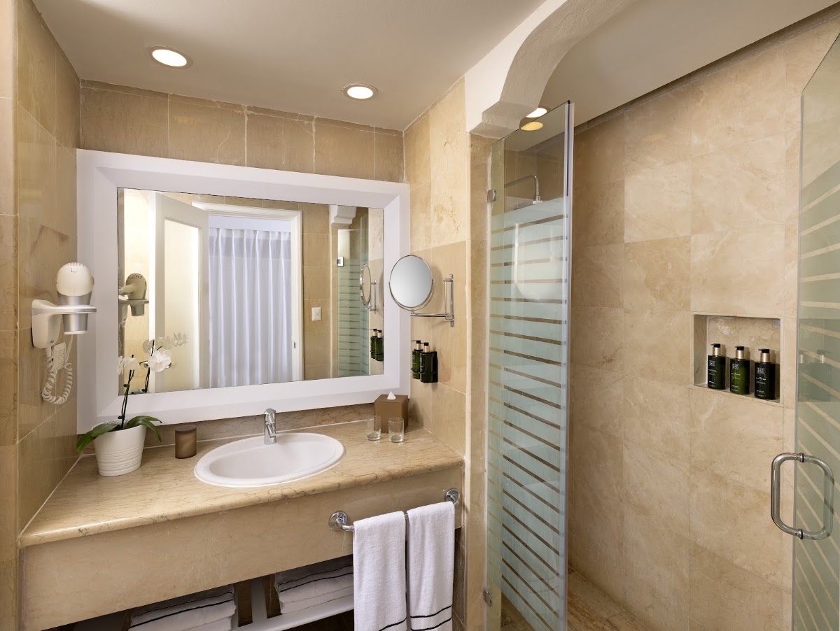Bathroom with cream marble walls, illuminated mirror above curved sink, and glass-enclosed shower with heated towel rack.
