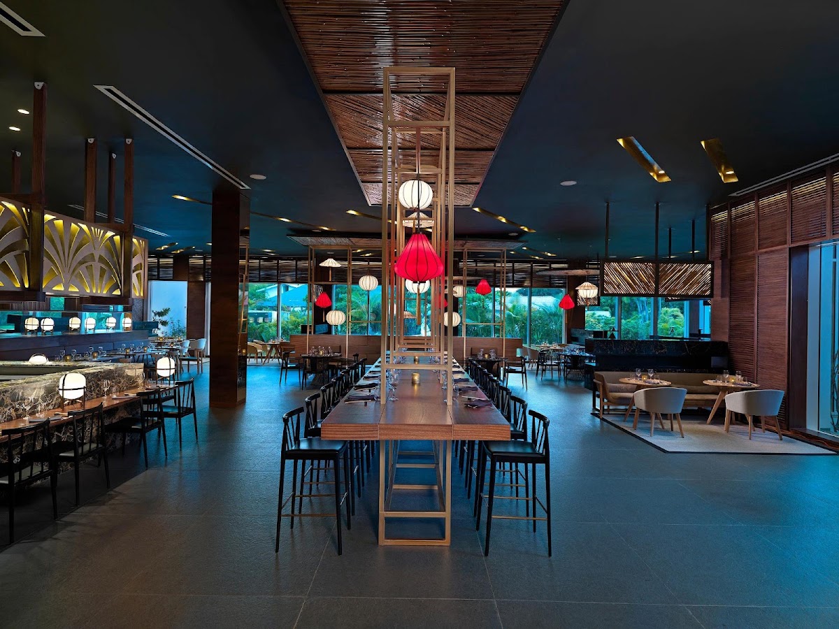 Modern dining room with long wood table, black chairs, red paper lanterns, and floor-to-ceiling windows overlooking tropical grounds at dusk.