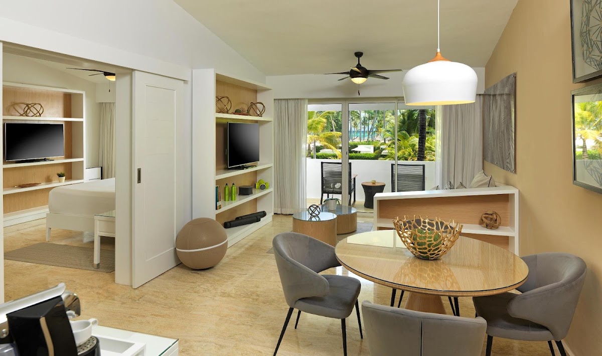 Modern resort suite with dining table, gray chairs, wood furnishings, TV, and glass doors opening to tropical grounds at Meliá Punta Cana Beach Wellness Inclusive.