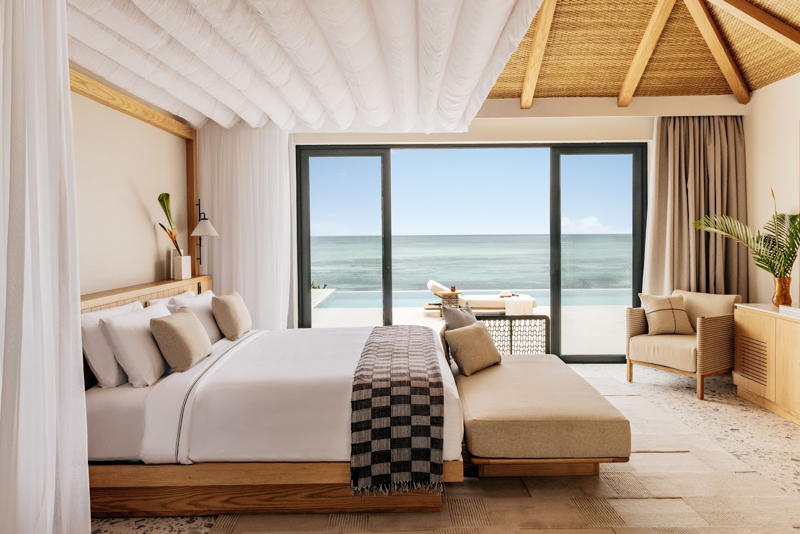 Beachfront bedroom at Meliá Zanzibar with ocean views through glass doors, white ceiling, wooden beam details, and lounge seating.