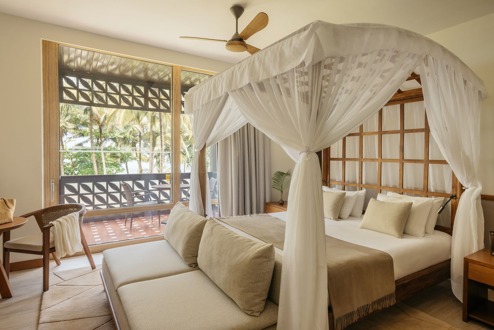 Spacious resort bedroom with a four-poster canopy bed draped in white curtains, sitting area, and ocean view balcony with palm trees visible outside.