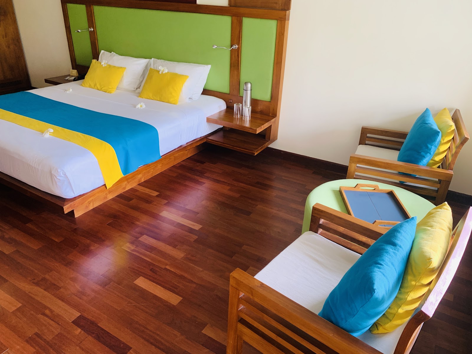 Guest room at Mermaid Hotel & Club with bed, wooden chairs, glass balcony door, and tropical view through windows.