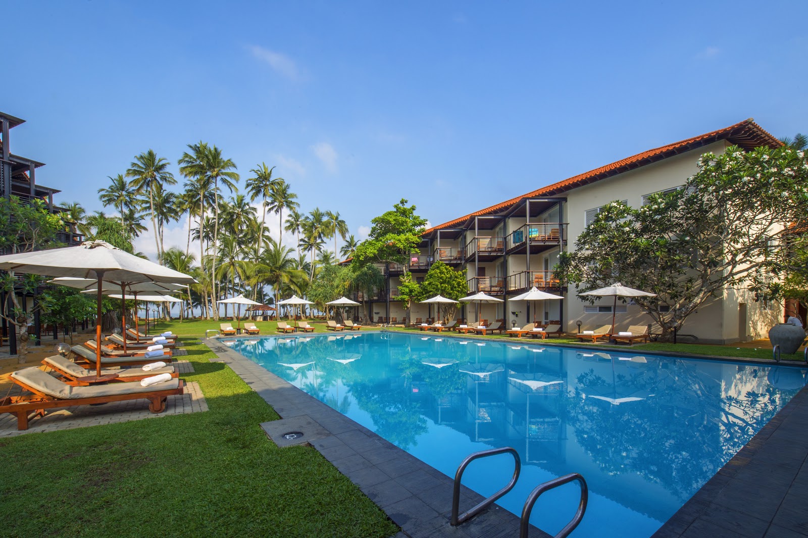 Resort pool with loungers and white umbrellas, flanked by manicured lawn and multi-story buildings with terracotta roofs, palm trees visible beyond.