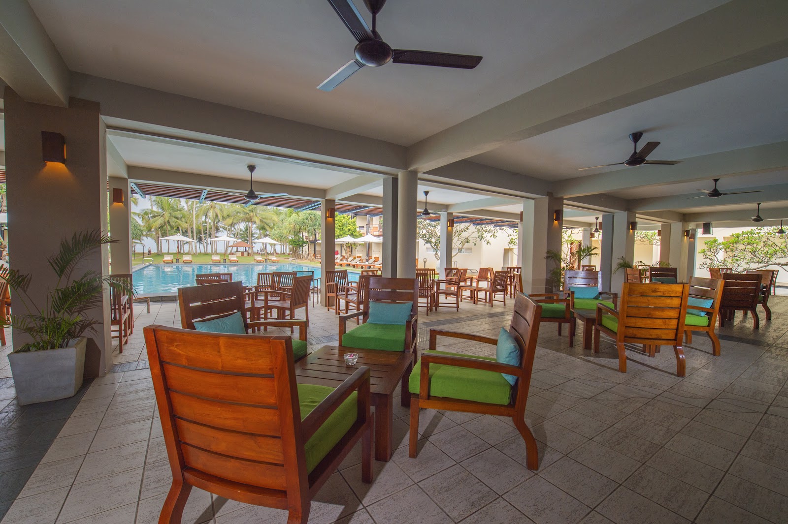 Covered lounge area with wooden chairs and green cushions overlooking a swimming pool and palm trees at Mermaid Hotel & Club.