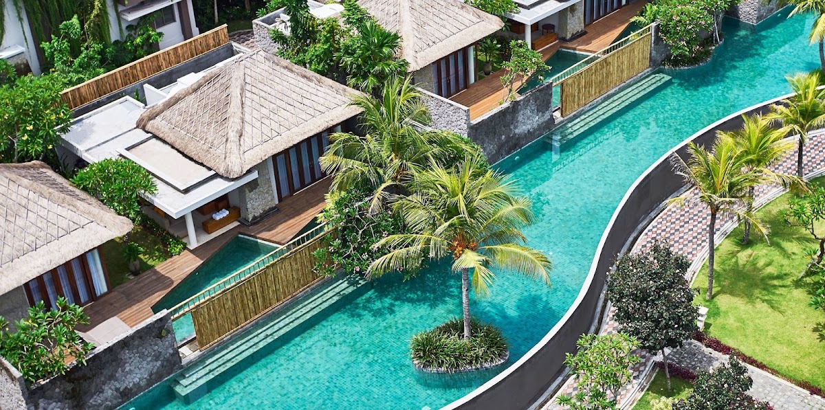 Aerial view of resort villas with thatched roofs surrounding a curved turquoise pool lined with palm trees and tropical landscaping.