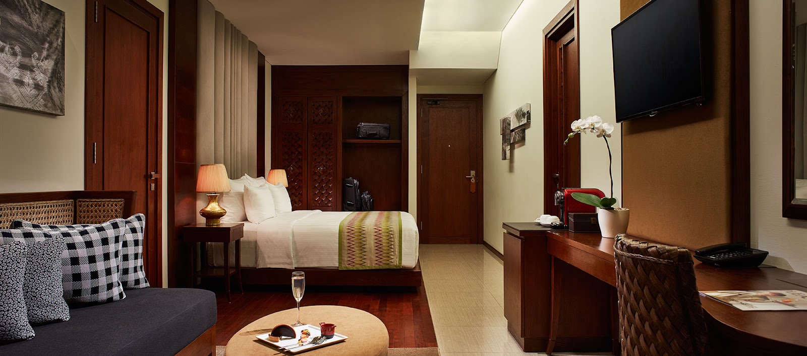 Hotel room with dark wood doors and furnishings, queen bed with white linens, sofa with patterned pillows, desk with orchid plant, and flat-screen TV.