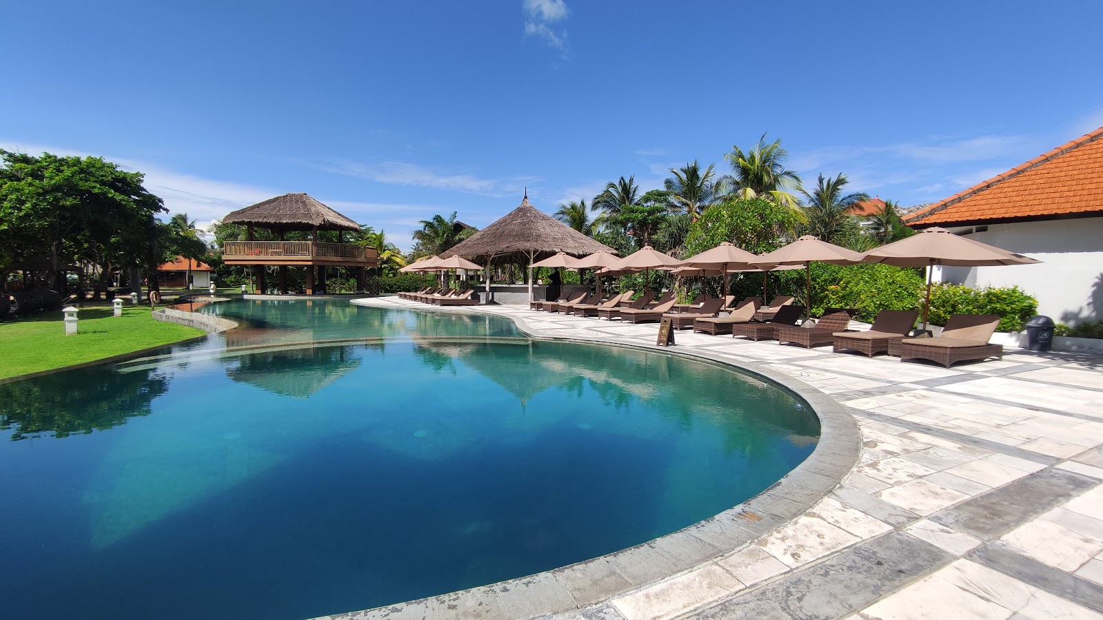 Curved turquoise pool with stone deck, lounge chairs under tan umbrellas, thatched-roof buildings, palm trees, and manicured lawn at Merusaka Nusa Dua.