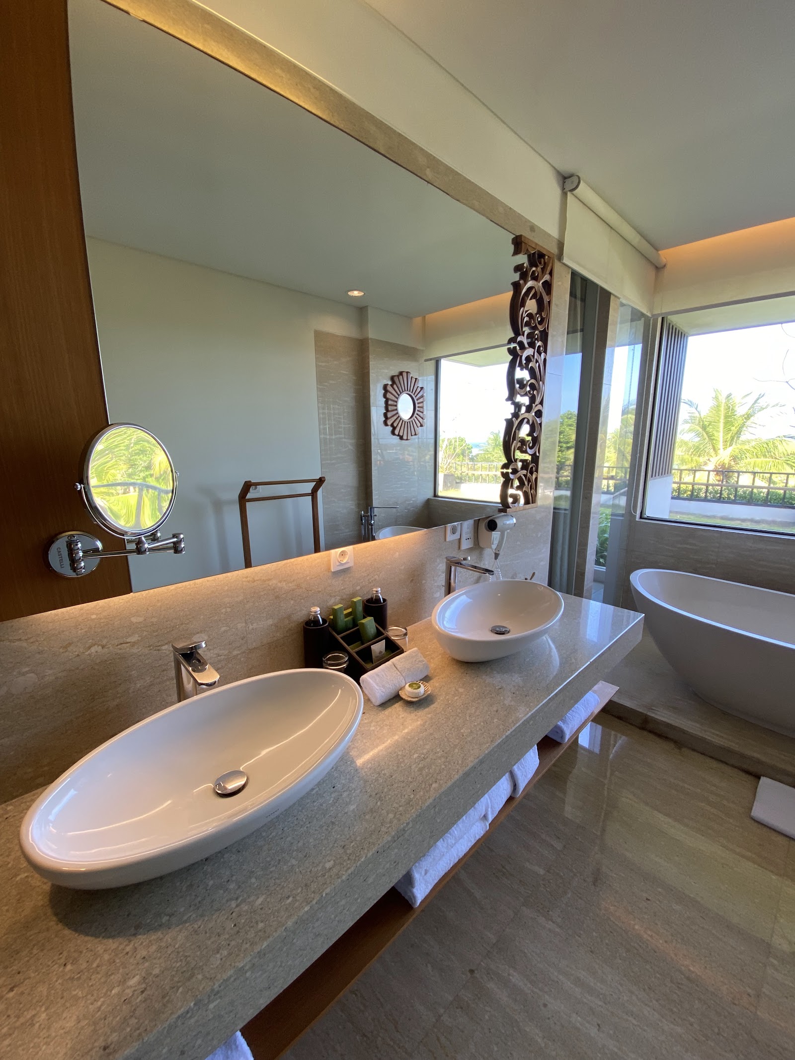 Modern bathroom at Merusaka Nusa Dua with dual sinks, large mirror, soaking tub, and ocean view through floor-to-ceiling windows.