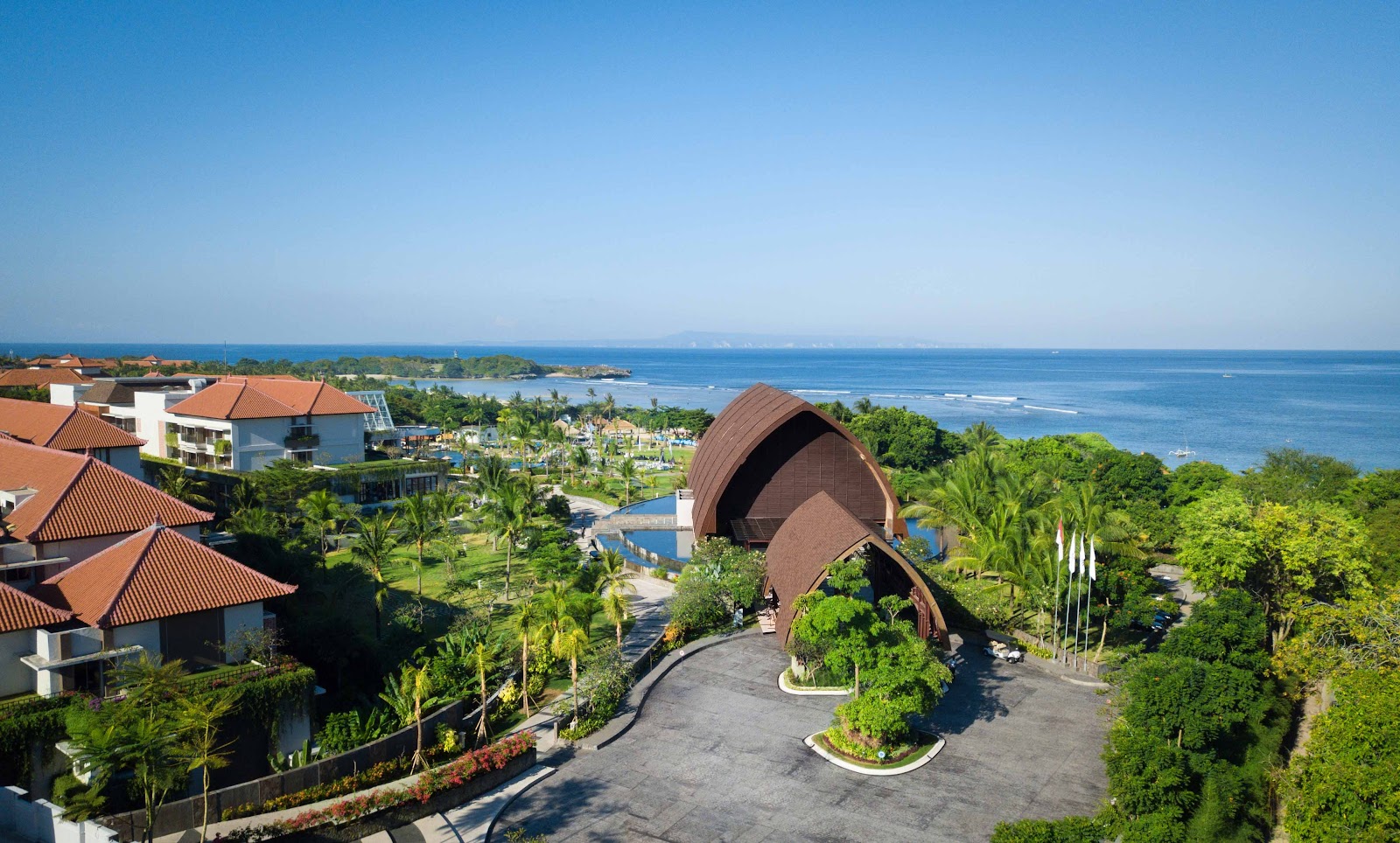 Aerial view of Merusaka Nusa Dua with curved architecture, terracotta roofs, tropical grounds, pools, and ocean beyond.