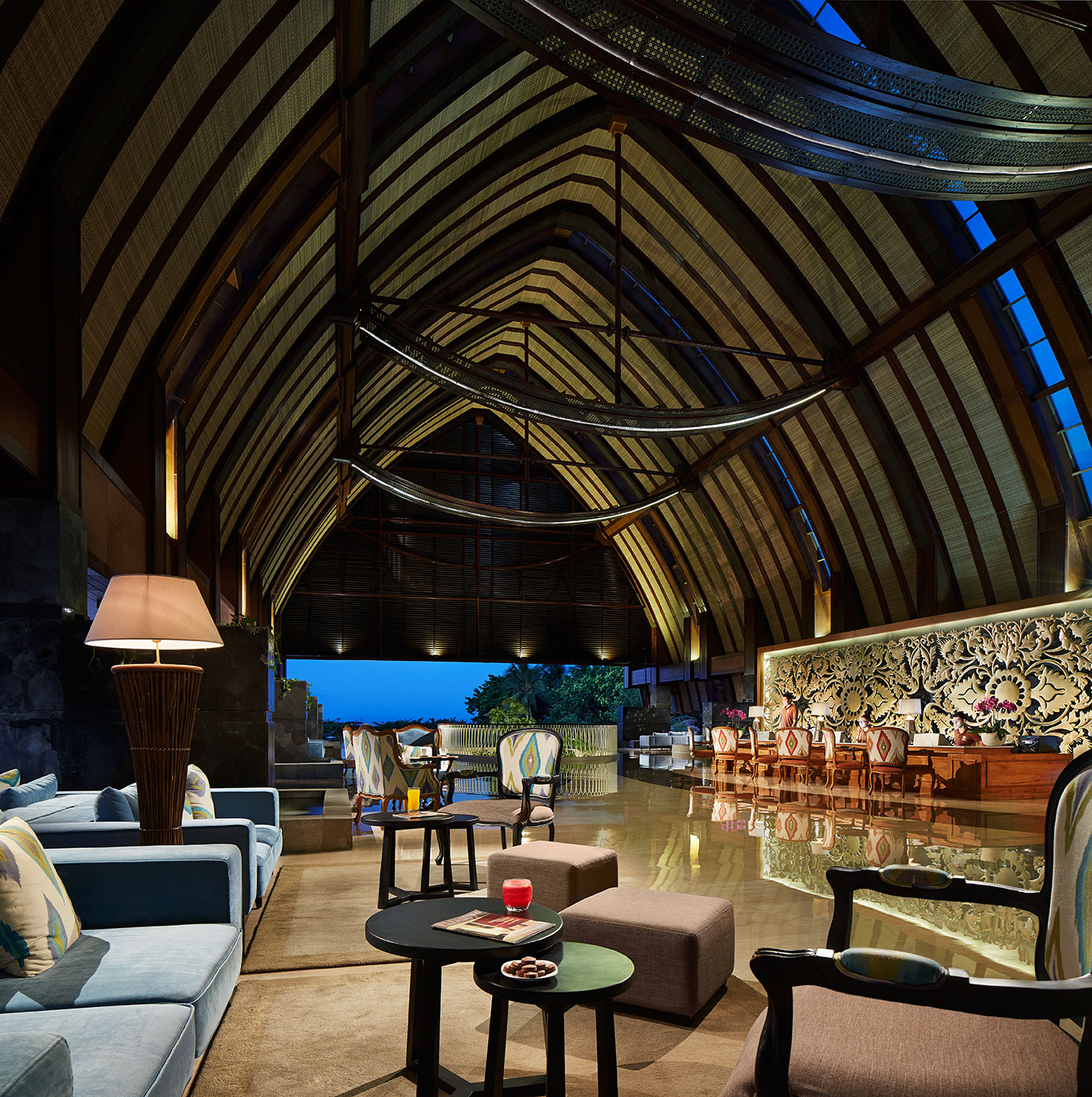 Merusaka Nusa Dua's soaring open-air lounge features dramatic arched wooden beams, contemporary furnishings, and a carved stone accent wall overlooking a twilight terrace.