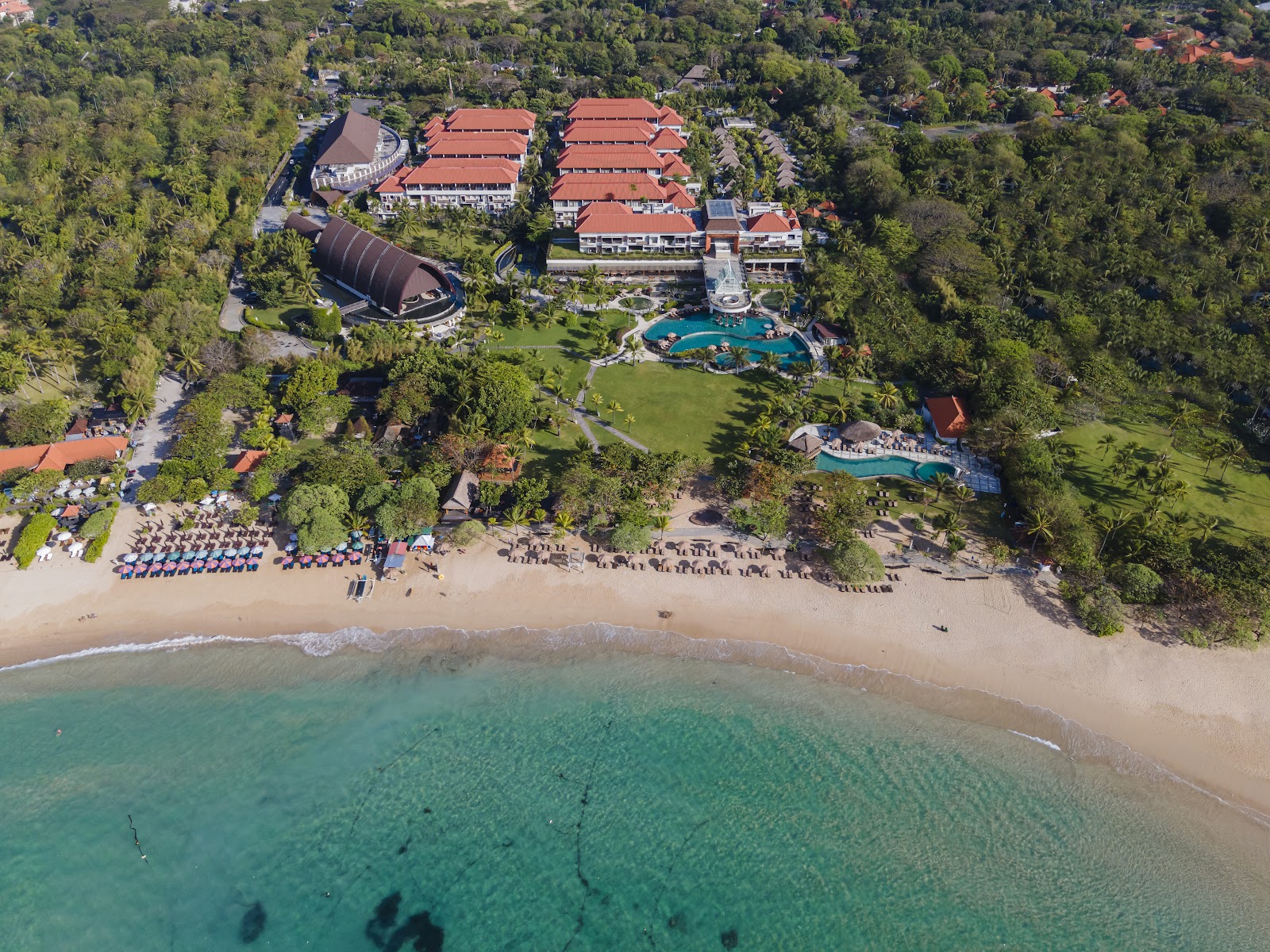 Aerial view of resort with red-roofed buildings, lagoon pool, and beach lined with blue umbrellas along turquoise water.