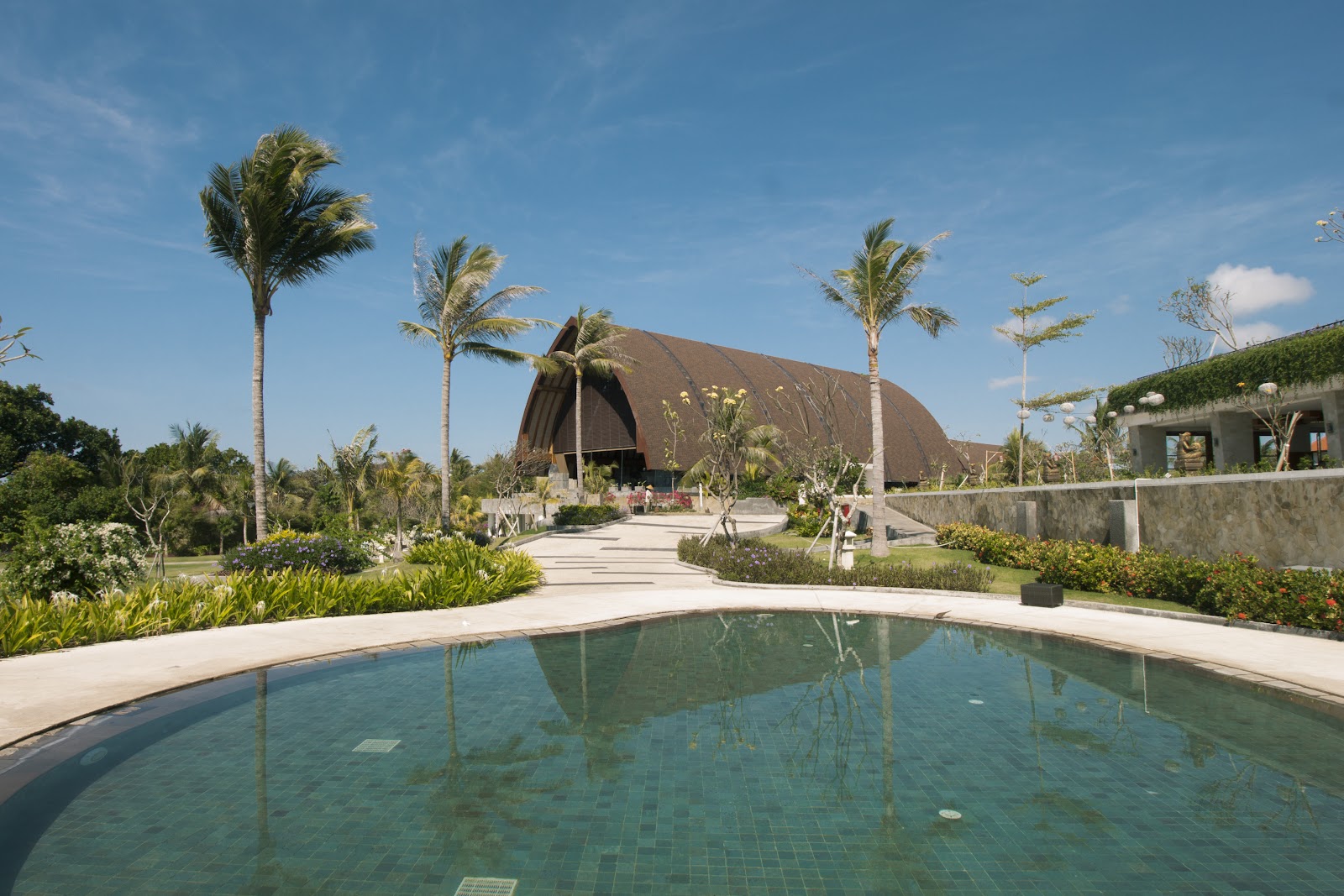 Curved pool with tiled floor reflects palm trees and a large dome-shaped building with dark roof at Merusaka Nusa Dua.