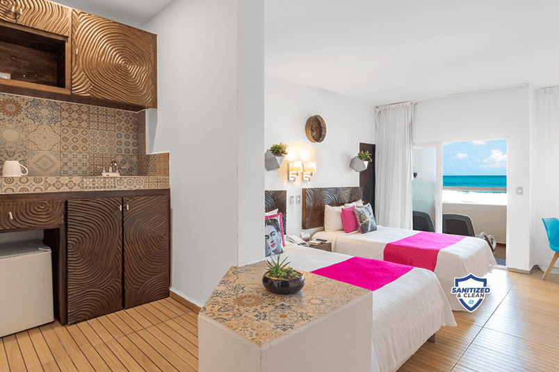 Modern resort room with two beds topped in magenta, kitchenette with patterned backsplash, and ocean view through glass doors at MIA Reef Isla Mujeres.