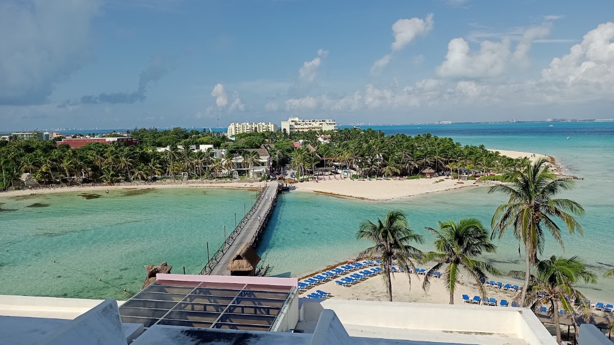 MIA Reef Isla Mujeres overlooks a turquoise lagoon with a long pier, white sand beach lined with blue loungers, and palm trees under clear sky.
