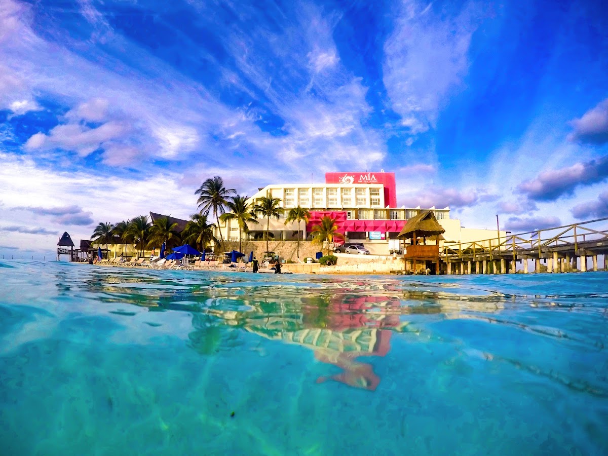Clear turquoise water in the foreground with the MIA Reef Isla Mujeres resort visible across the beach, featuring a pink-signed building and palm trees under blue sky.