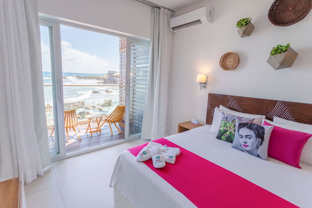 Oceanfront bedroom at MIA Reef Isla Mujeres with bed, balcony seating, and views of rocky coastline and turquoise water.