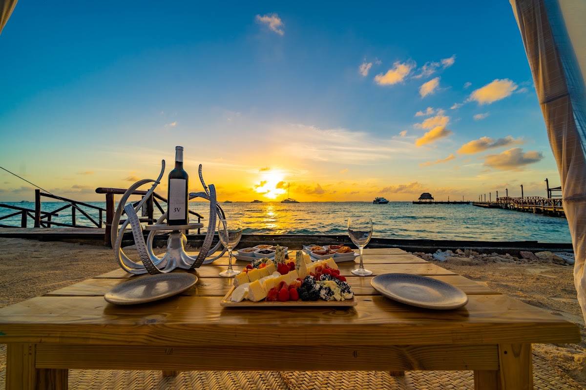 Beachfront dinner table at sunset with wine bottle, place settings, and fruit platter overlooking the ocean and wooden dock.