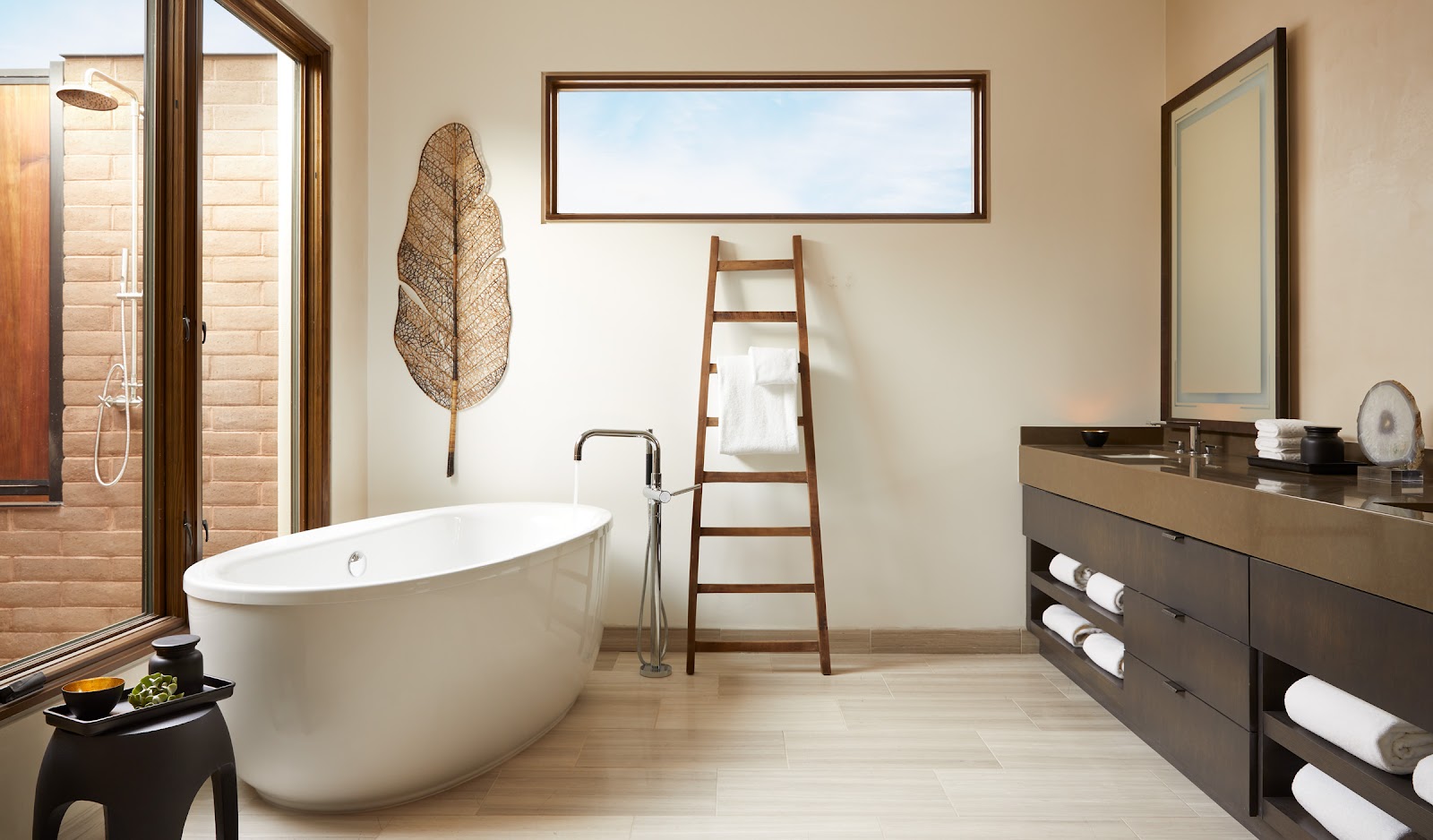 Modern bathroom at Miraval Arizona Resort & Spa with freestanding white tub, wooden ladder, woven leaf decor, and dark vanity with towel storage.