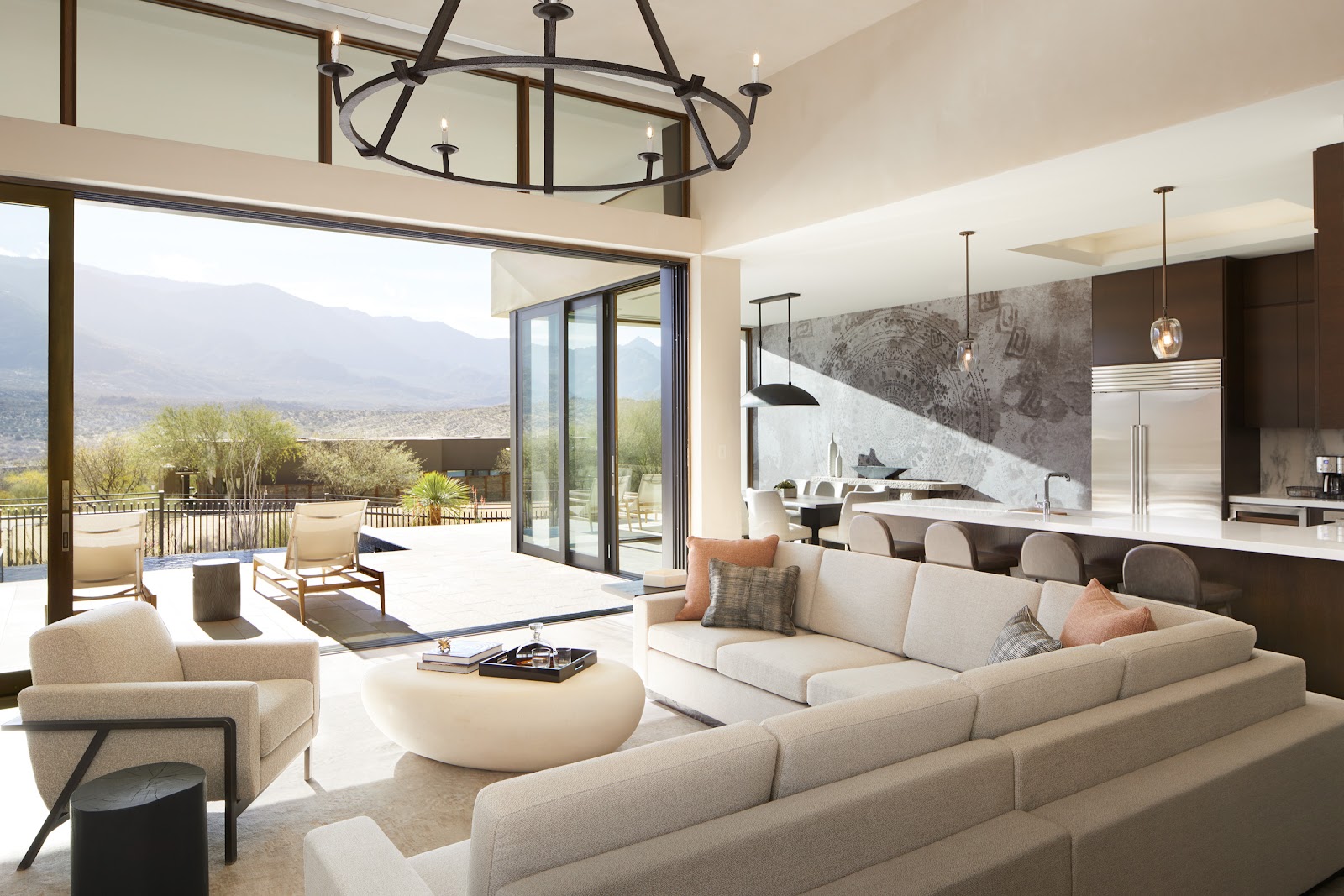 Open-concept suite with cream sectional sofa, modern kitchen, and floor-to-ceiling windows overlooking desert mountains and landscaped terrace.