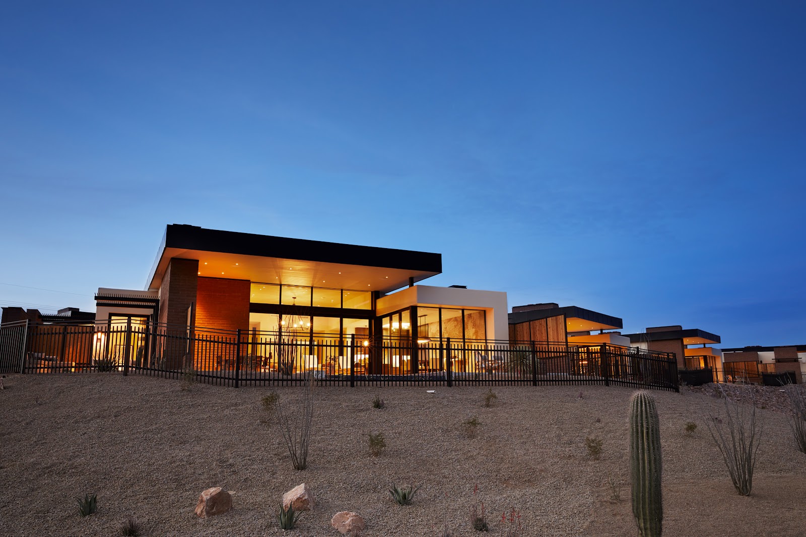 Modern resort villas with orange and black accents sit on desert landscaping with saguaro cactus and rocks under clear blue sky at dusk.
