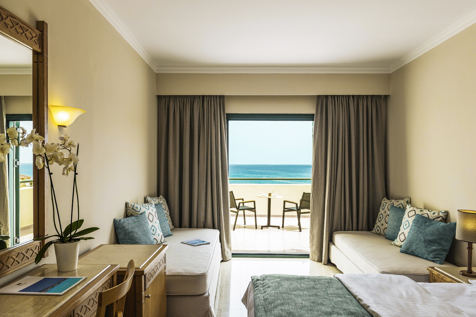 Resort room with two beds, cream walls, and open doors leading to a beachfront balcony overlooking the ocean and sandy beach.