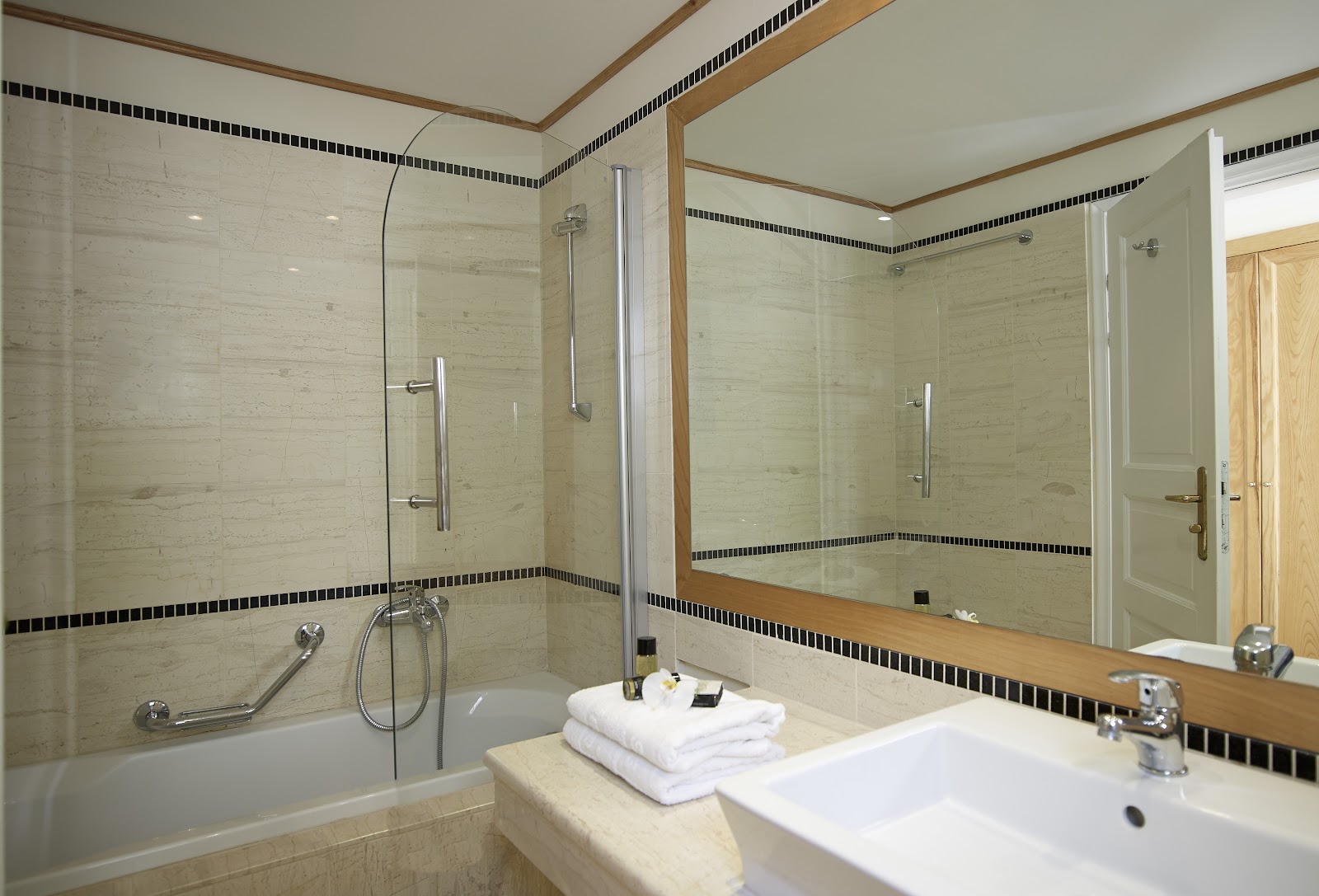 Bathroom with cream travertine tile, black tile border stripe, glass shower enclosure, soaking tub, double vanity with white sink, and folded white towels on the counter.