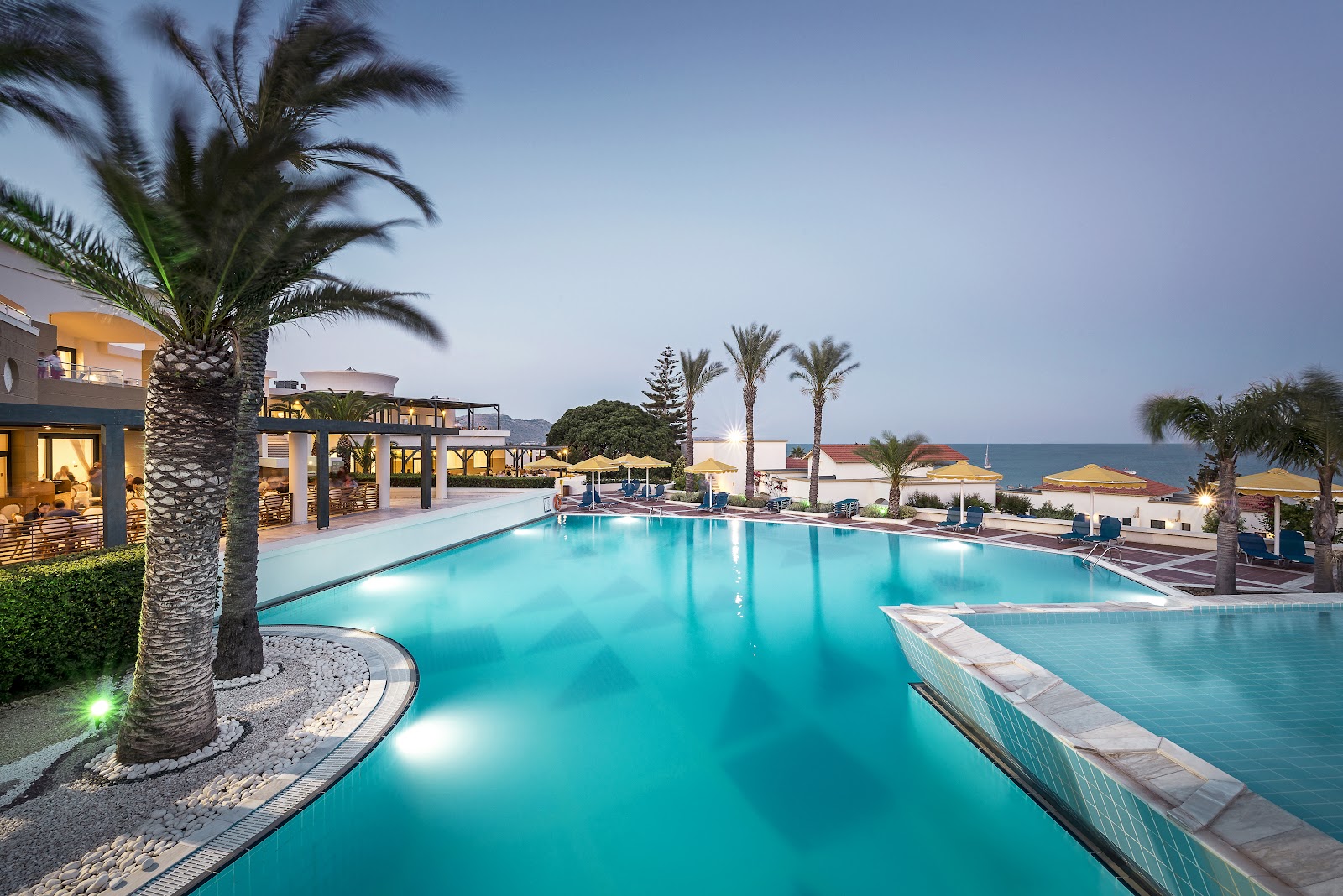 Two plated desserts and iced beverages sit on a white poolside ledge overlooking turquoise water at Mitsis Rodos Maris.