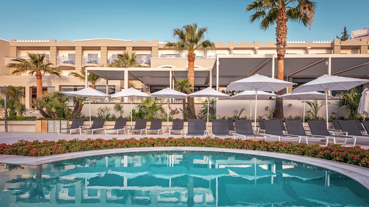 Curved pool with turquoise water bordered by loungers, white umbrellas, and flowering beds, with a beige resort building and palm trees beyond.