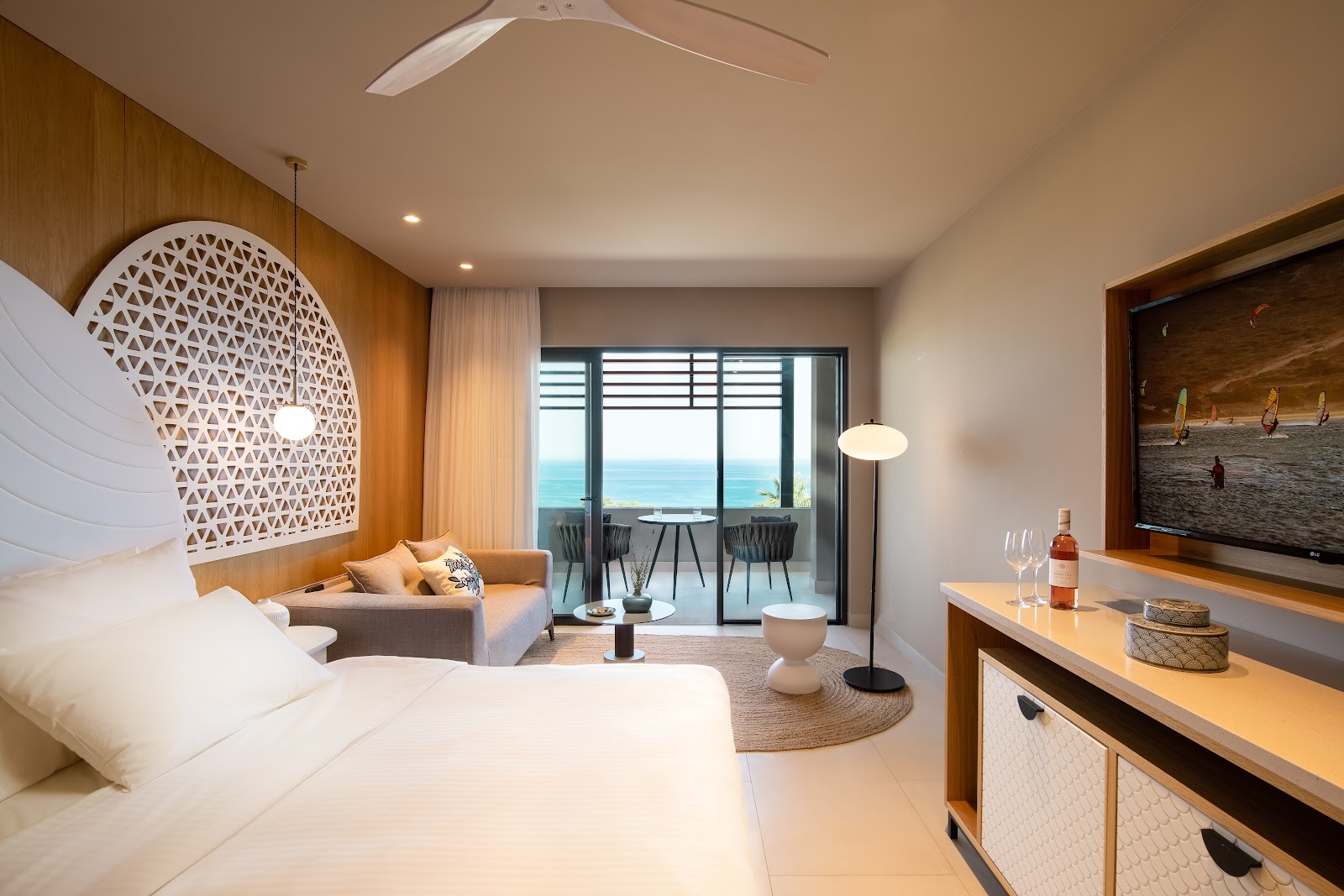 Resort suite with ocean view from balcony, featuring bed with geometric headboard, seating area, and modern wood furnishings overlooking turquoise sea.