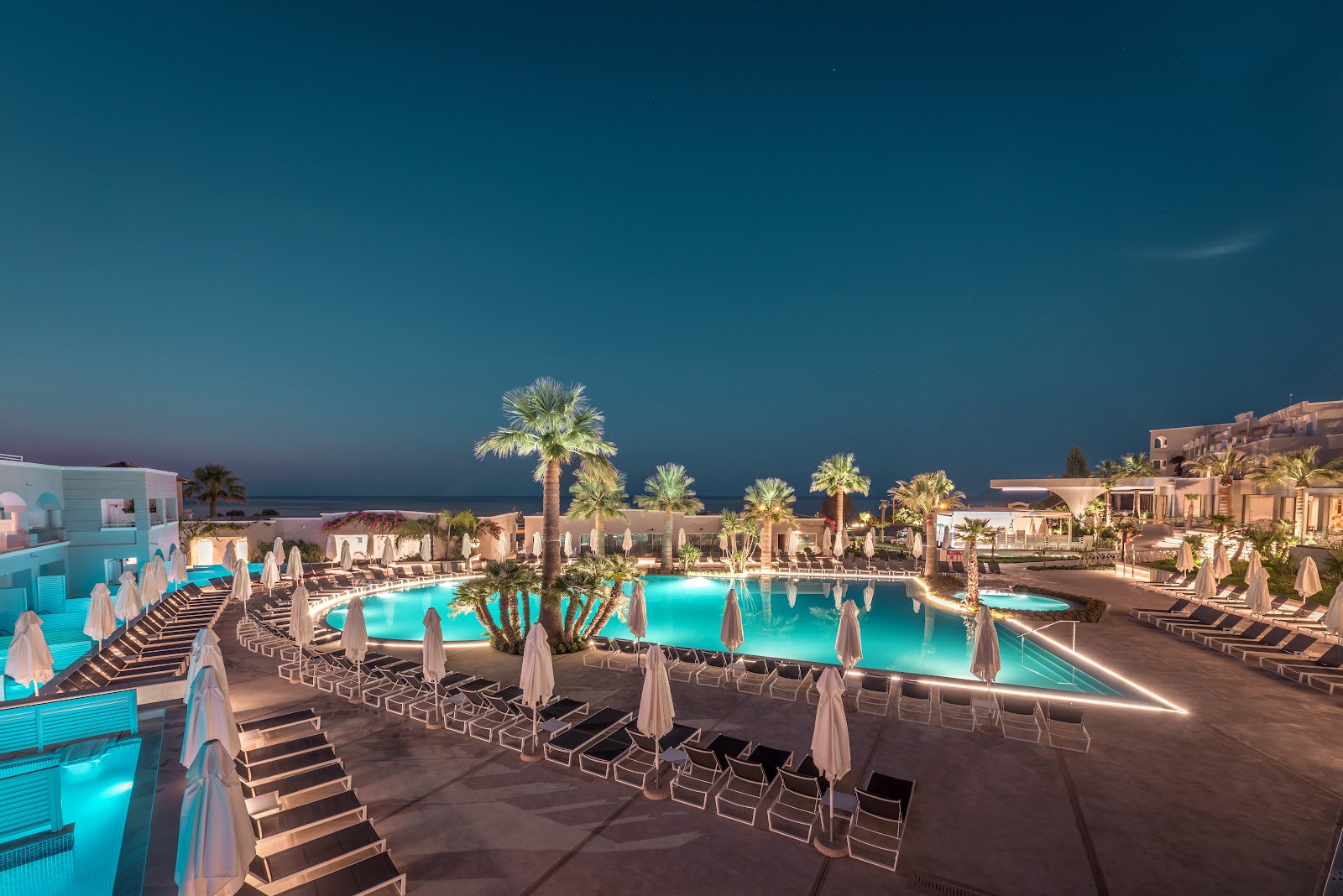 Twilight view of Mitsis Rodos Village with illuminated turquoise pools, white umbrellas, loungers, and palm trees overlooking the sea.