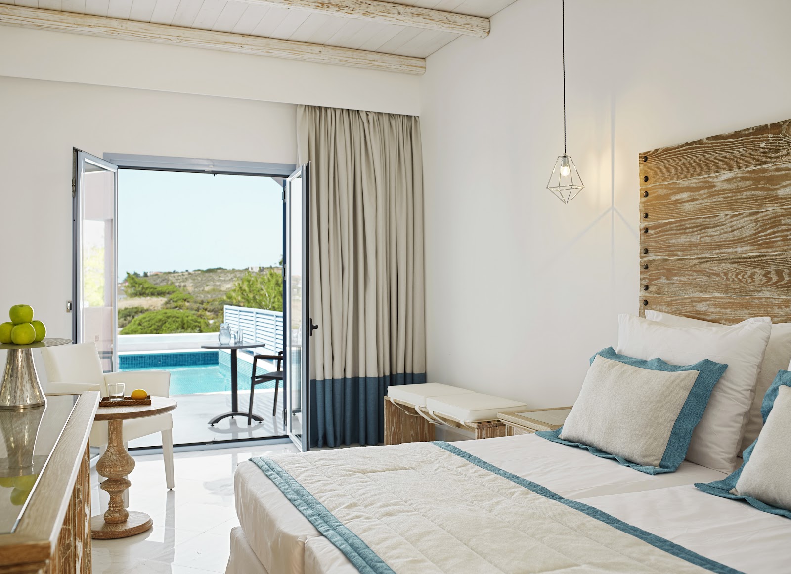 Resort bedroom with bed, wooden headboard, and open glass doors leading to a private pool overlooking Mediterranean hillside vegetation.