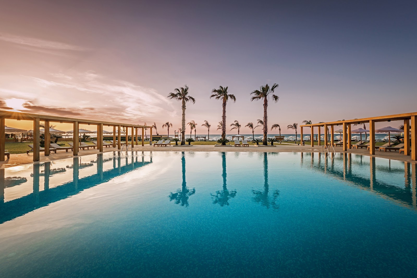 Covered pool with turquoise water reflecting palm trees and wooden pergolas at sunset, ocean and islands visible beyond the grounds.