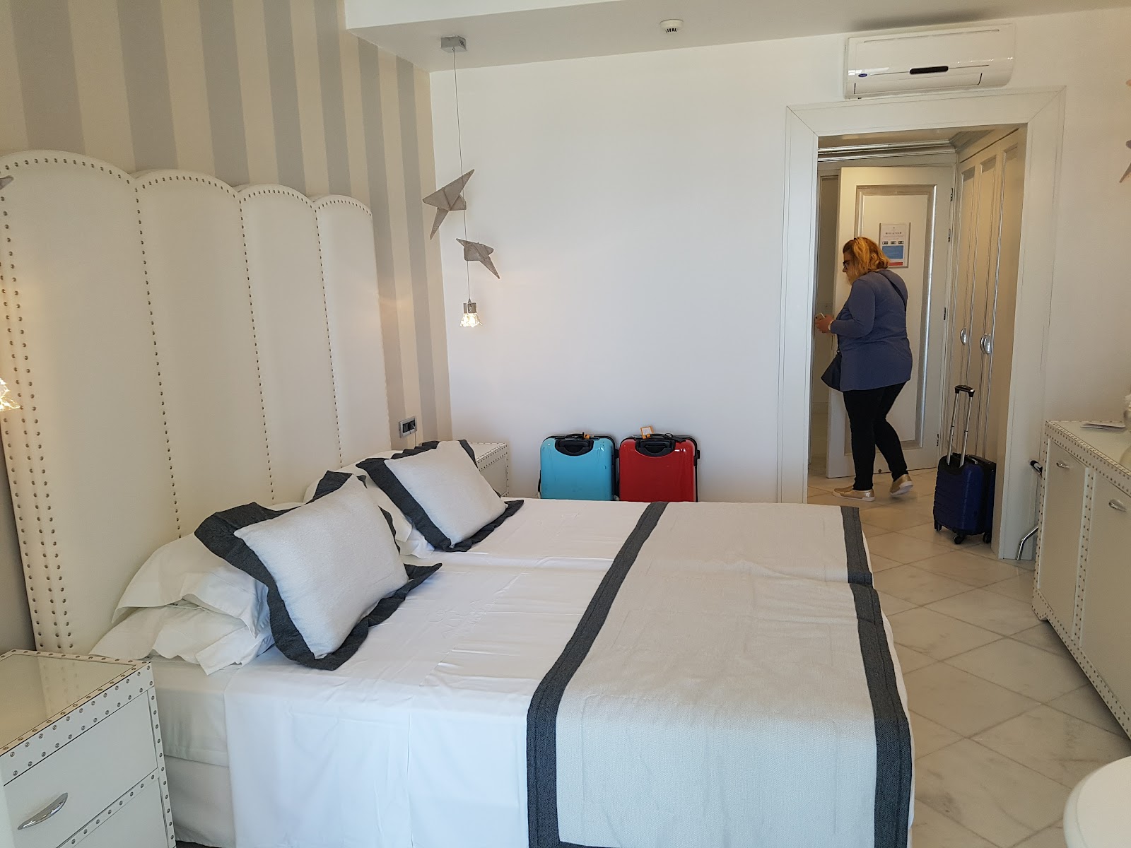 Resort room with queen bed, white upholstered headboard, blue and red luggage, and guest entering through doorway.