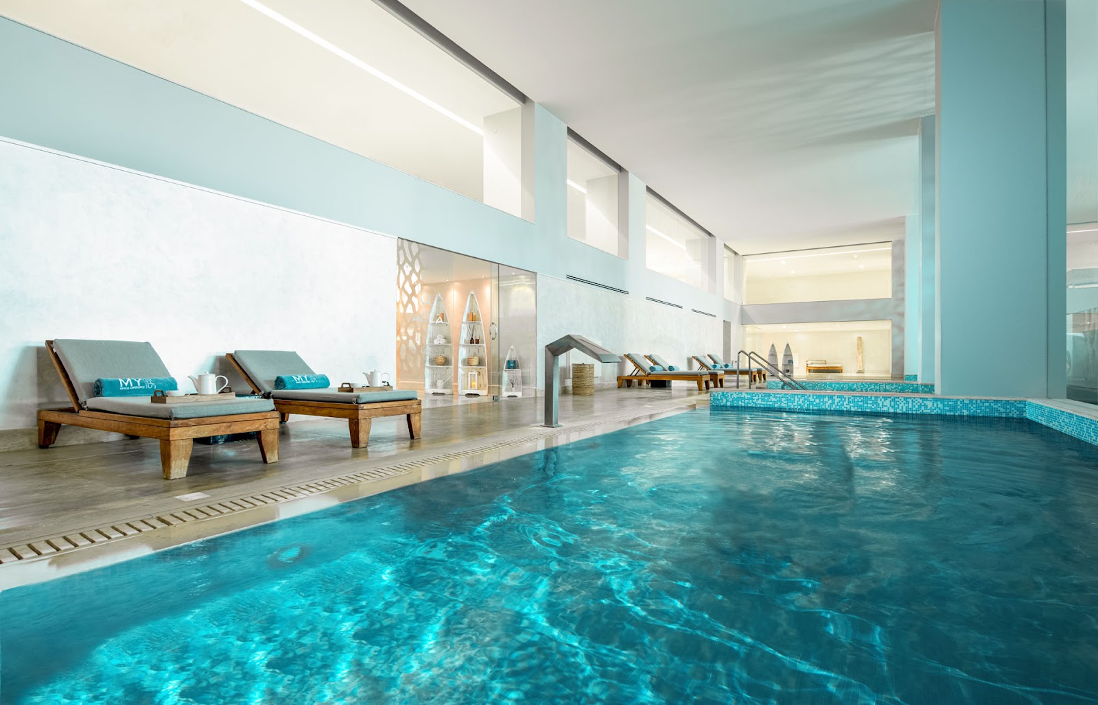 Indoor swimming pool with turquoise tile accents, wooden lounge chairs with cushions on the deck, and floor-to-ceiling windows overlooking a spa area.
