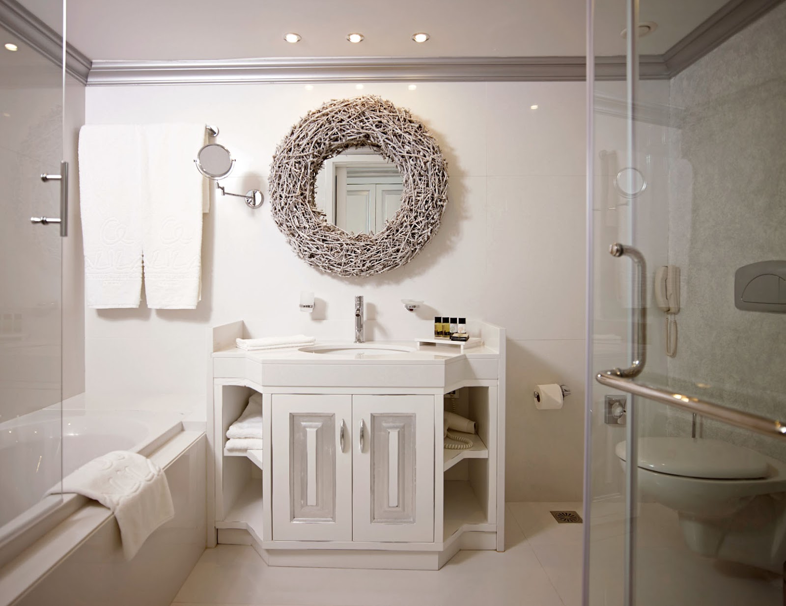 Modern bathroom with white vanity, round woven mirror, soaking tub, and glass-enclosed shower at Mitsis Selection Alila.