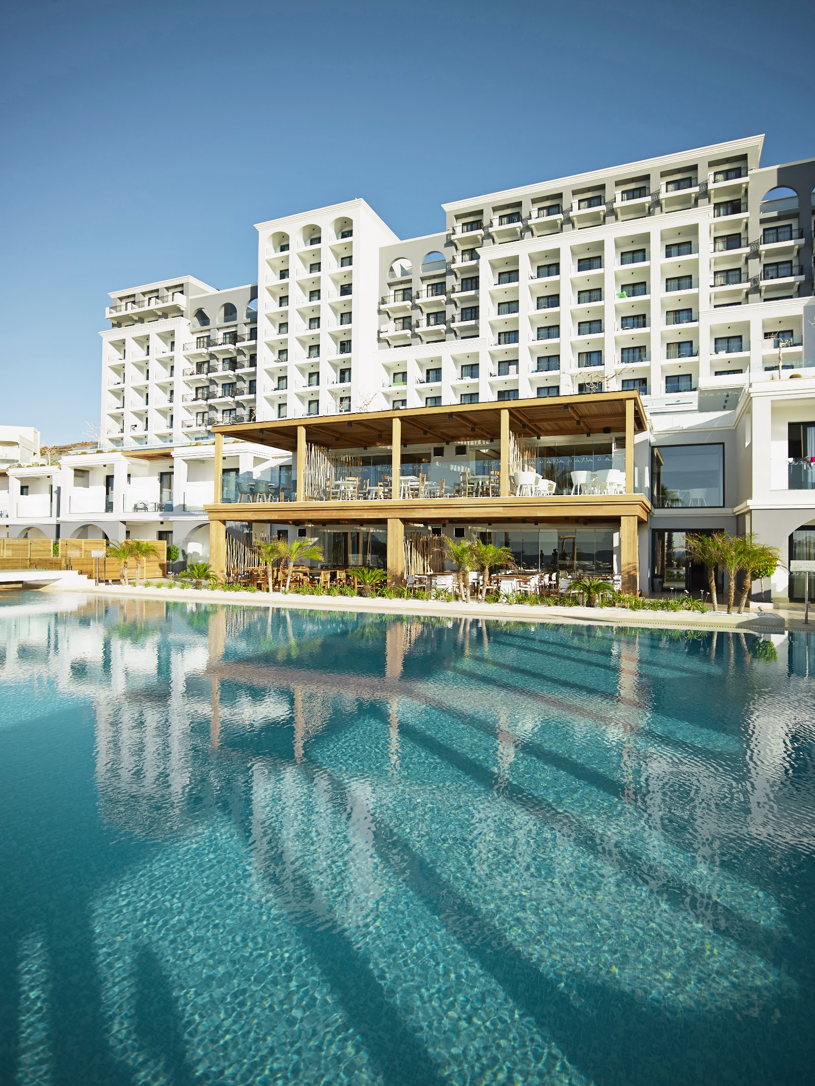 Resort pool with turquoise water reflecting white apartment buildings and wooden beachfront bar structure under clear blue sky.