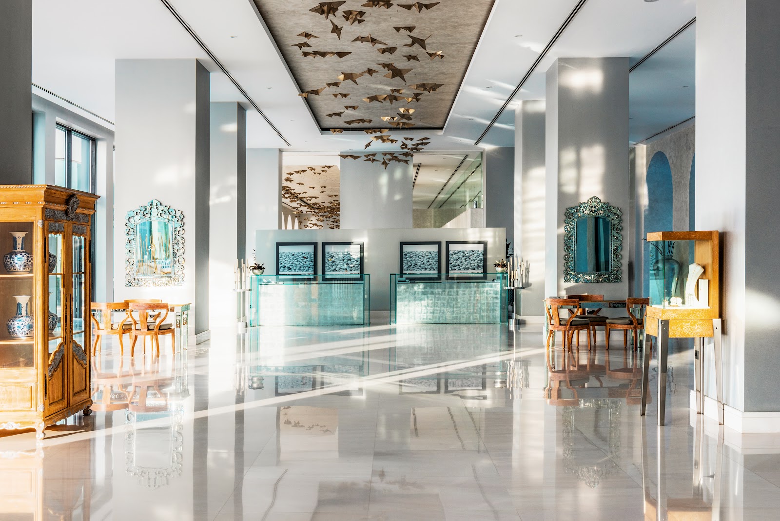 Modern resort lobby with polished white floor, ornate wooden furniture, turquoise-framed mirrors, and a ceiling decorated with bronze fish sculptures.