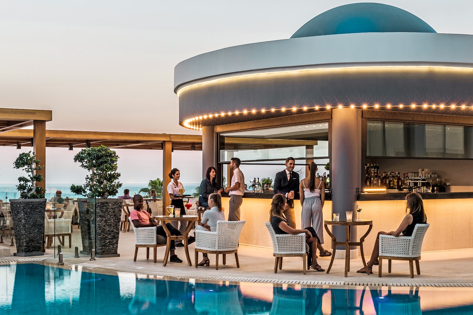 Guests relax in white chairs around an illuminated poolside bar at Mitsis Selection Alila, with the ocean visible beyond the terrace at dusk.