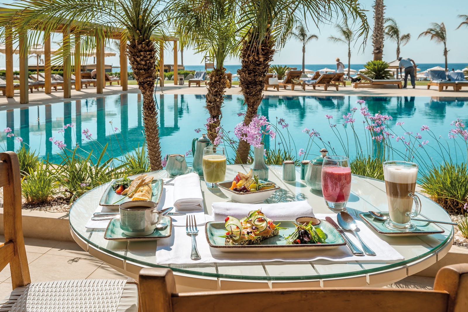 Beachfront dining table at Mitsis Selection Alila with plated food, beverages, and pink flowers overlooking a pool and Mediterranean Sea.