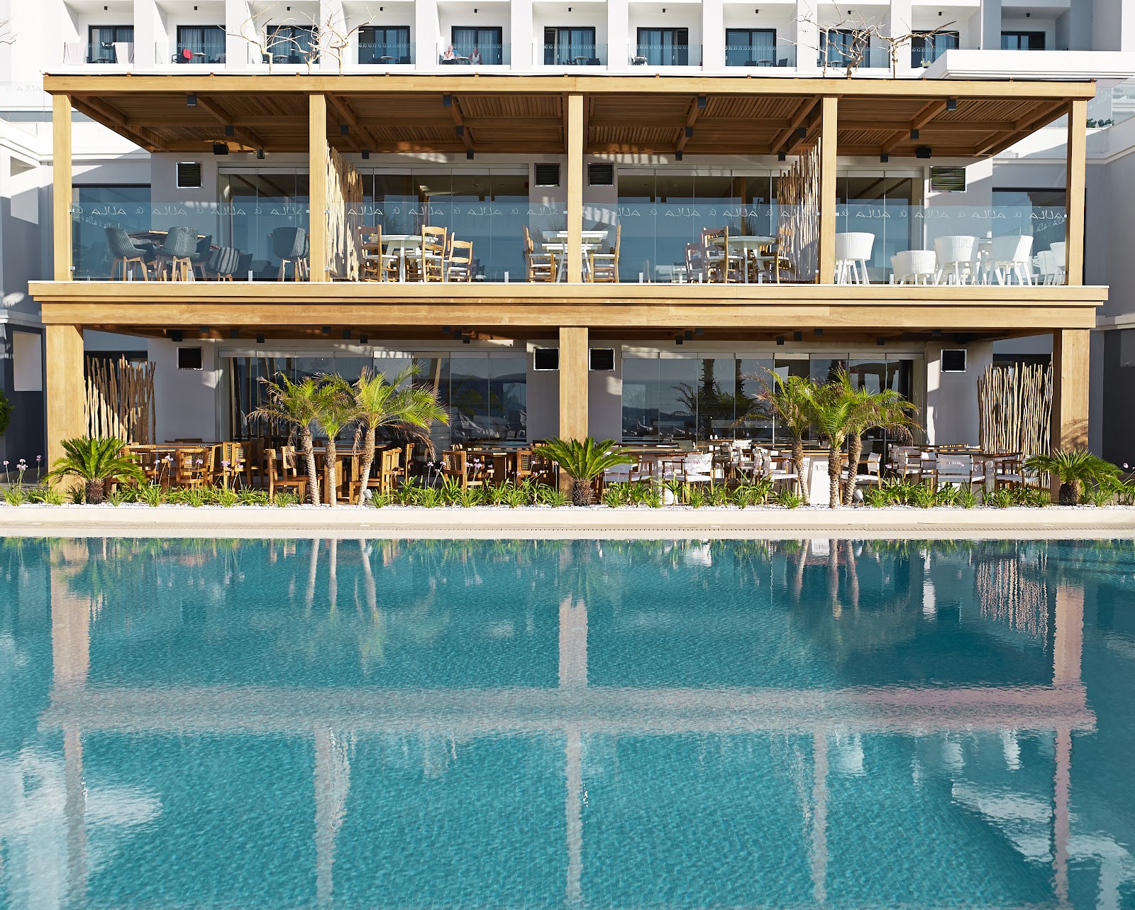 Turquoise pool with wooden pergola dining pavilion and palm plants along the edge, white resort building with guest rooms above.