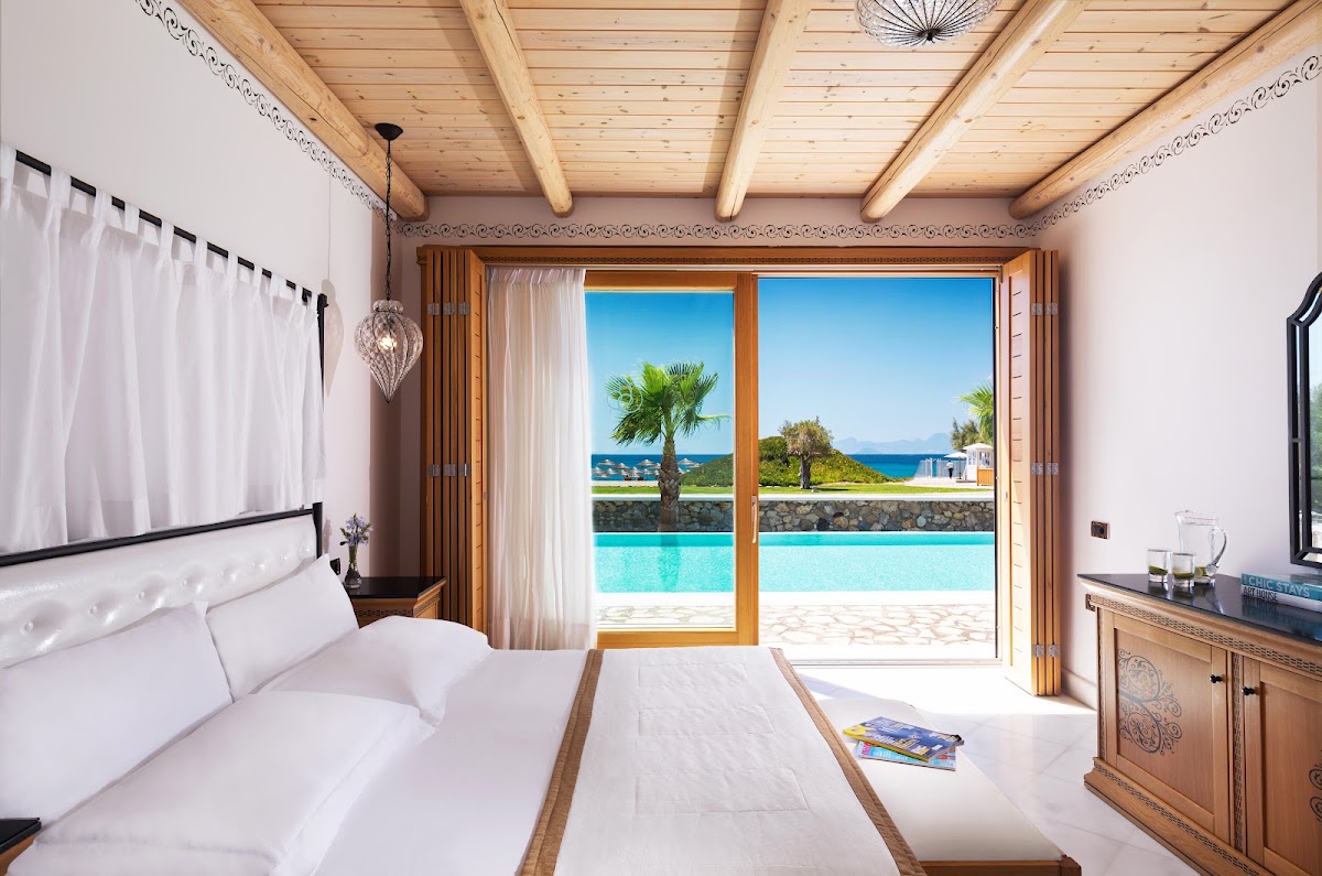 Bedroom with wooden beamed ceiling and white bedding overlooking a turquoise pool, palm trees, and blue sea at Mitsis Selection Blue Domes.