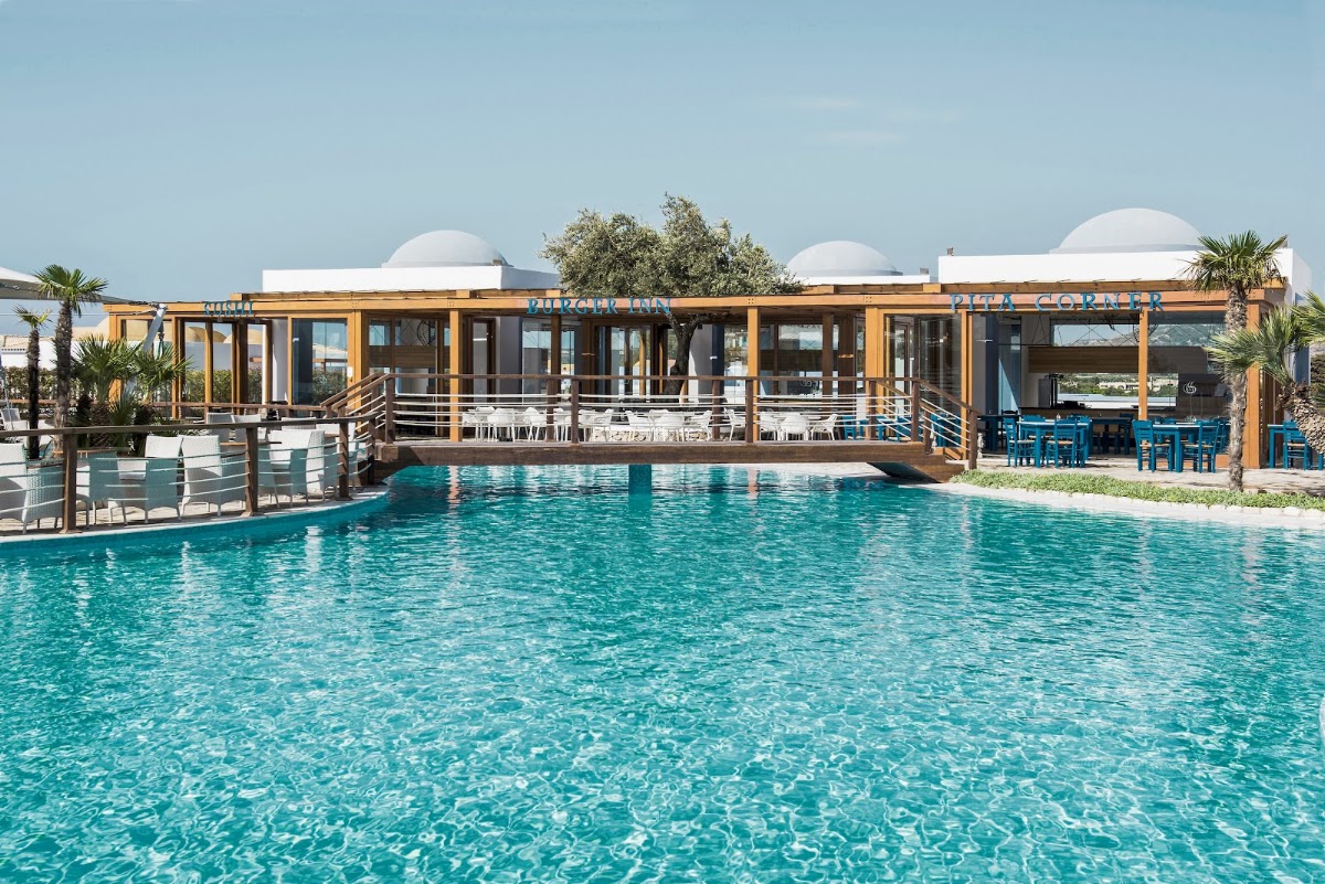 Turquoise pool facing a modern wooden pavilion with white dome structures, palm trees, and blue chairs at Mitsis Selection Blue Domes.